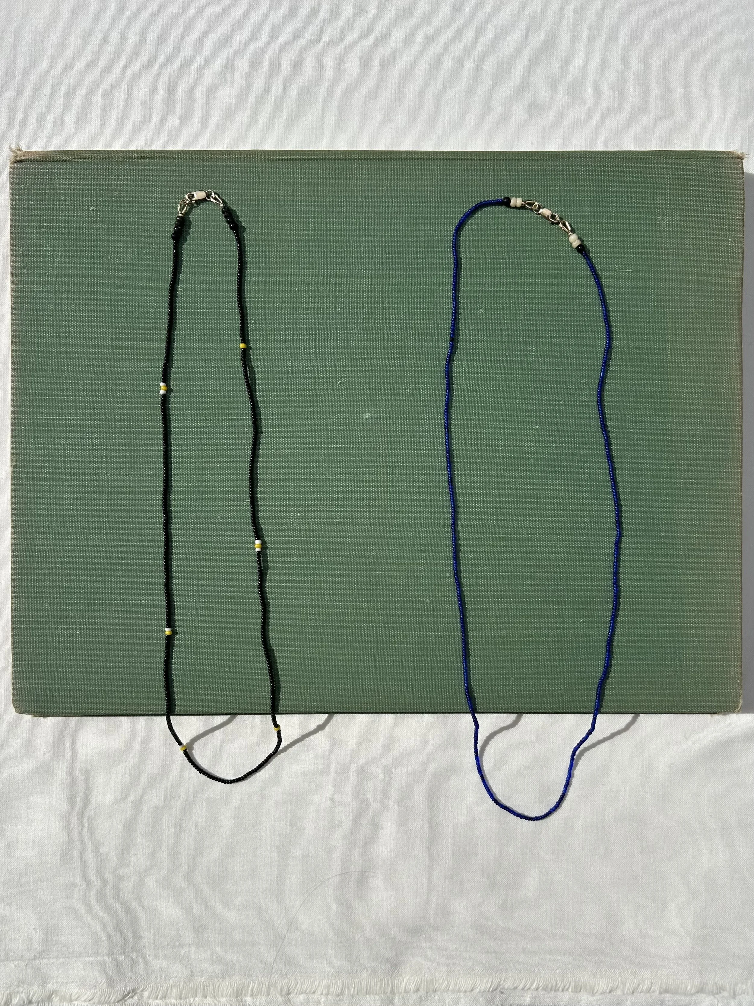 Two necklaces displayed on a green textured box; one black with yellow and white beads, one blue with white and black beads.