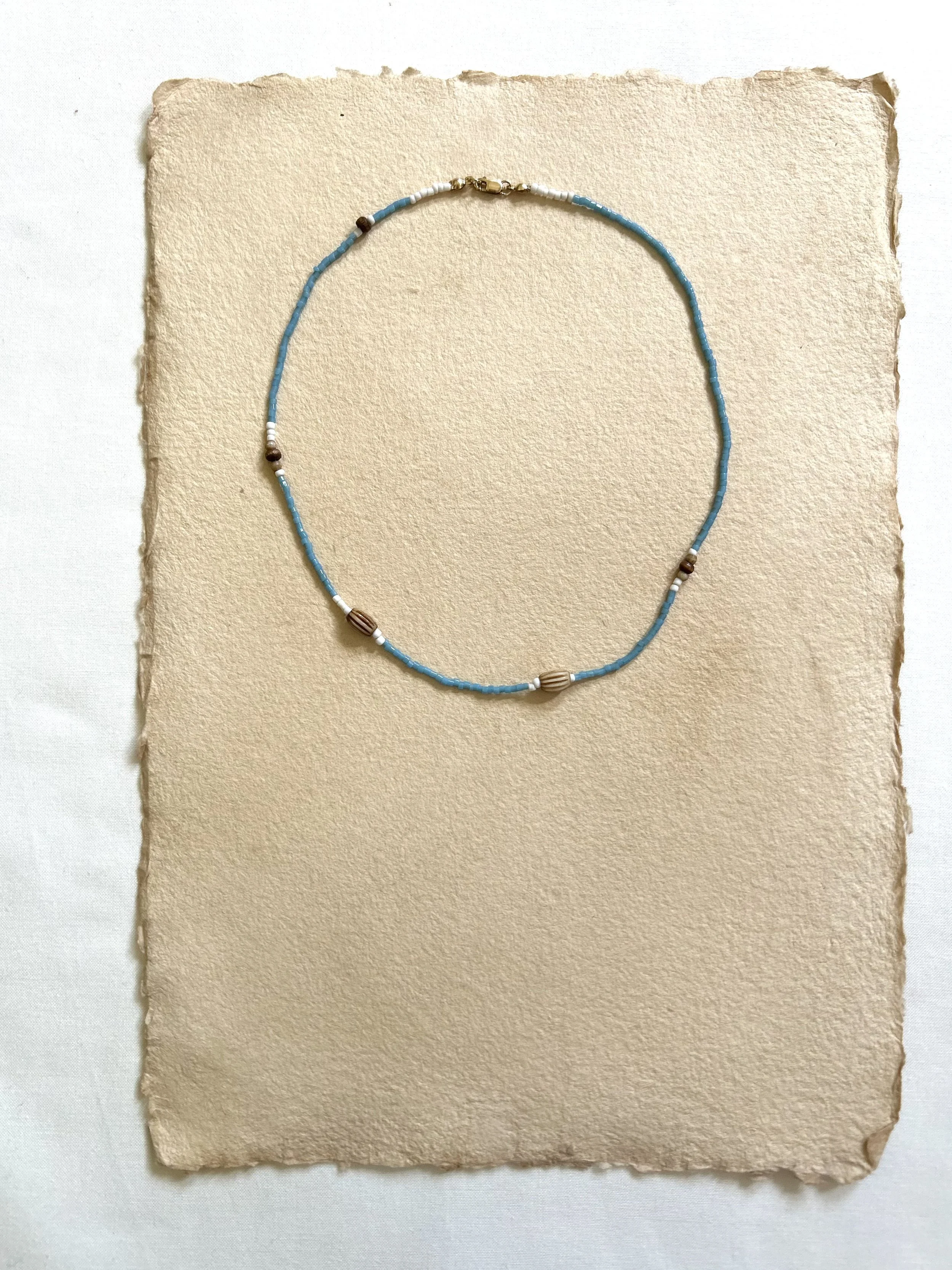 A beaded necklace with blue, white, brown, and beige beads displayed on a textured beige paper background.