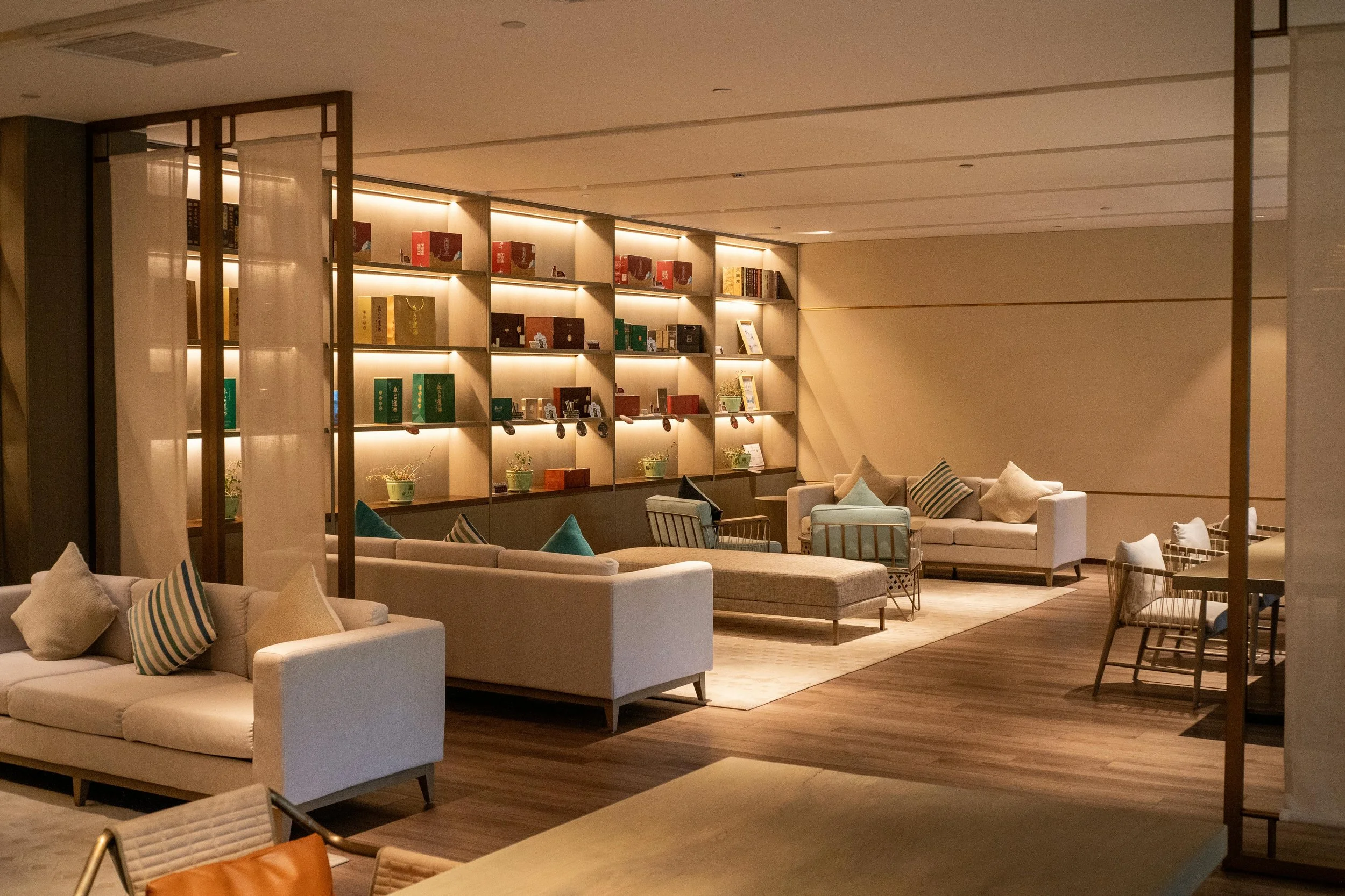A cozy, modern lounge area with beige sofas and striped pillows, wooden flooring, a large decorative shelving unit with books and plants, and soft large lighting.