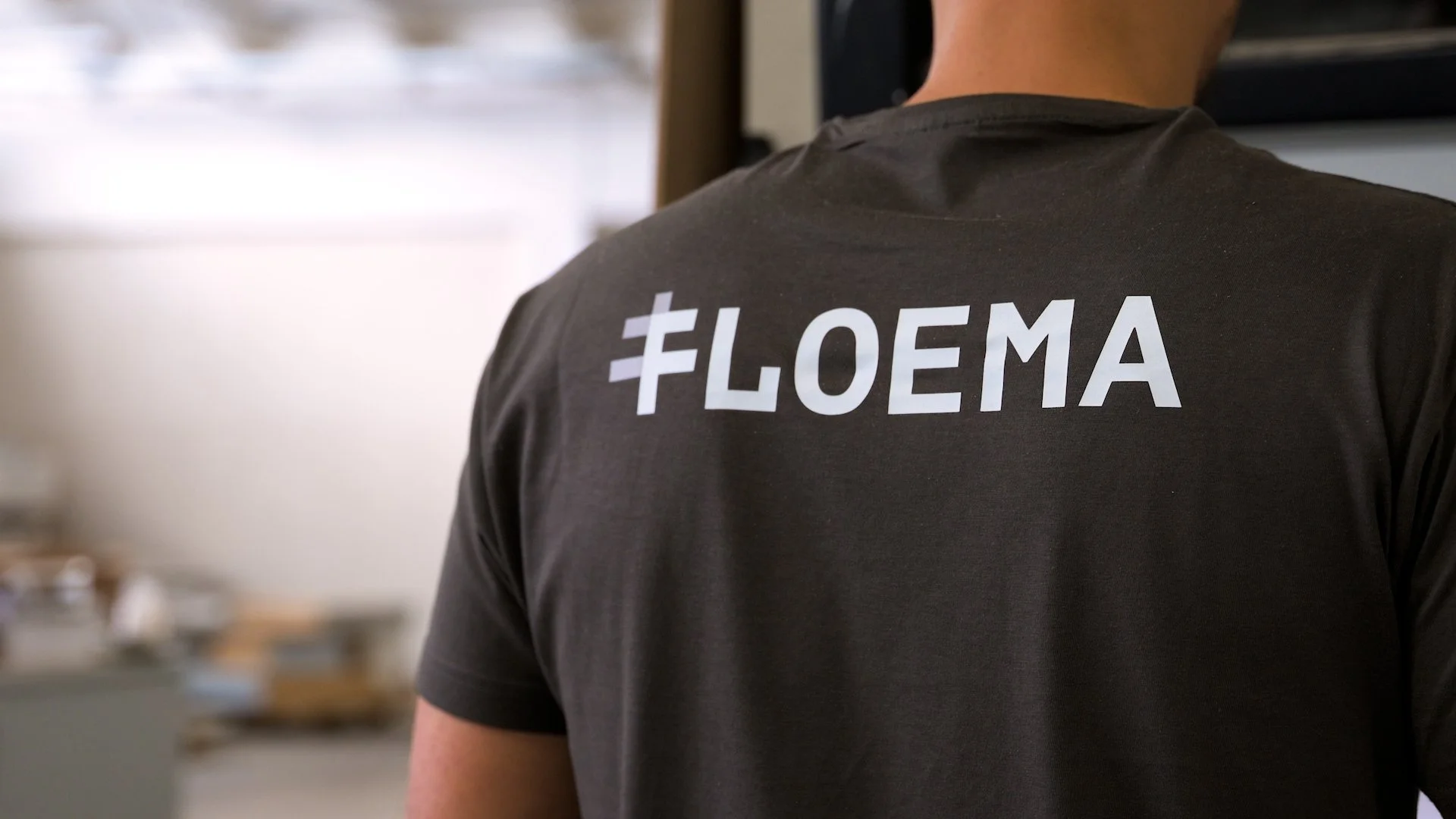 Person wearing a black T-shirt with the hashtag #FLOEMA printed on the back, standing indoors with a blurred background.