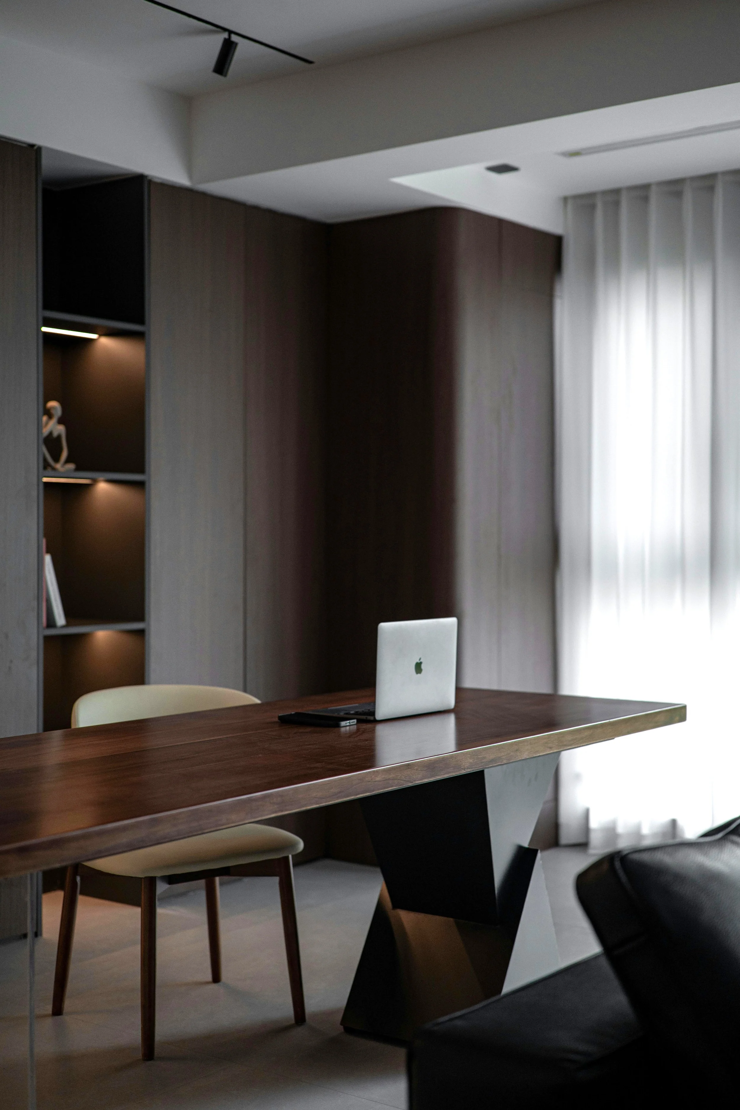 Modern office interior with a wooden table, a silver Apple laptop, beige chairs, dark furniture, and a curtain window.