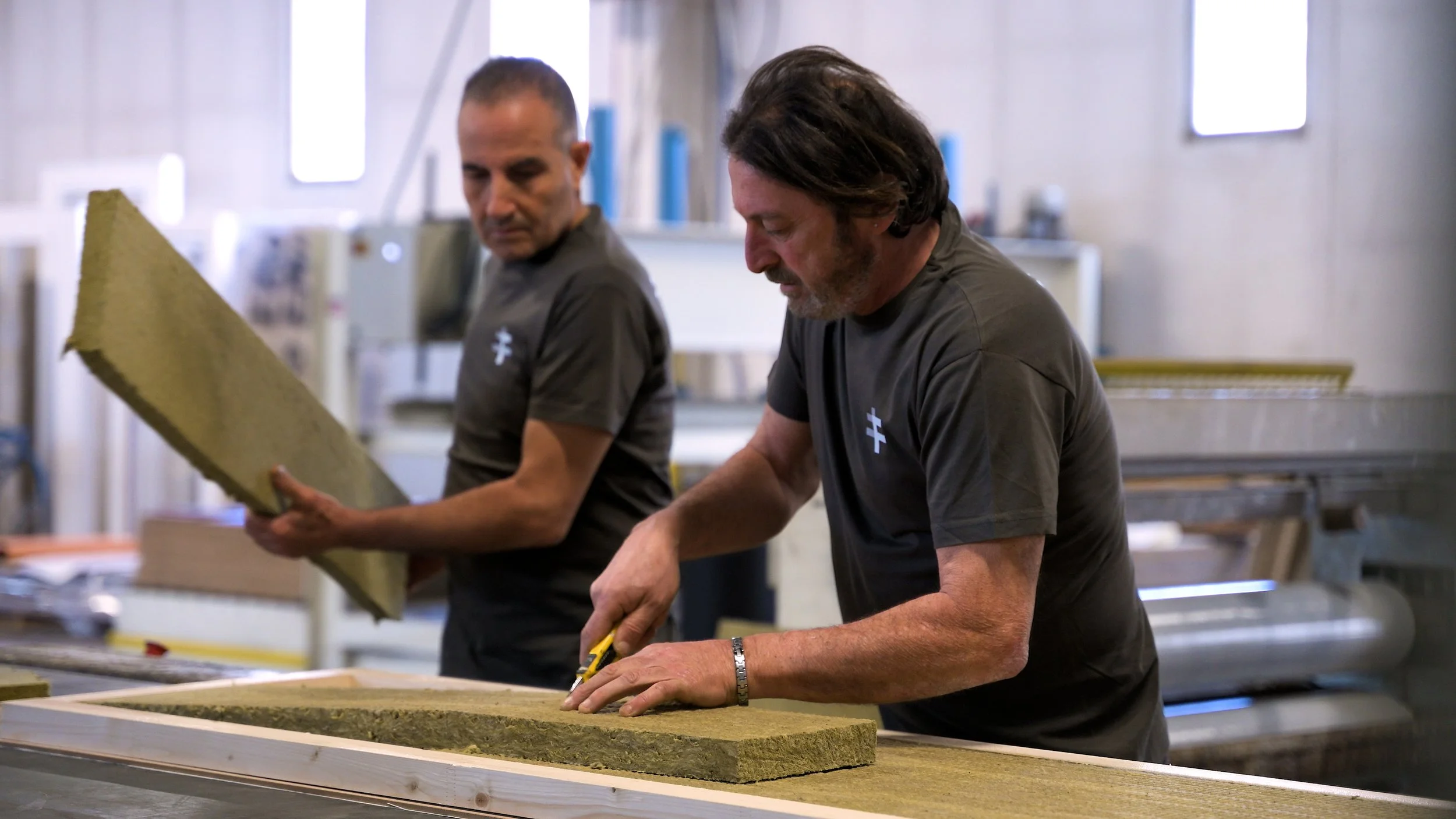 Two men in gray shirts working on a large flat surface, possibly crafting or manufacturing, in an industrial setting.