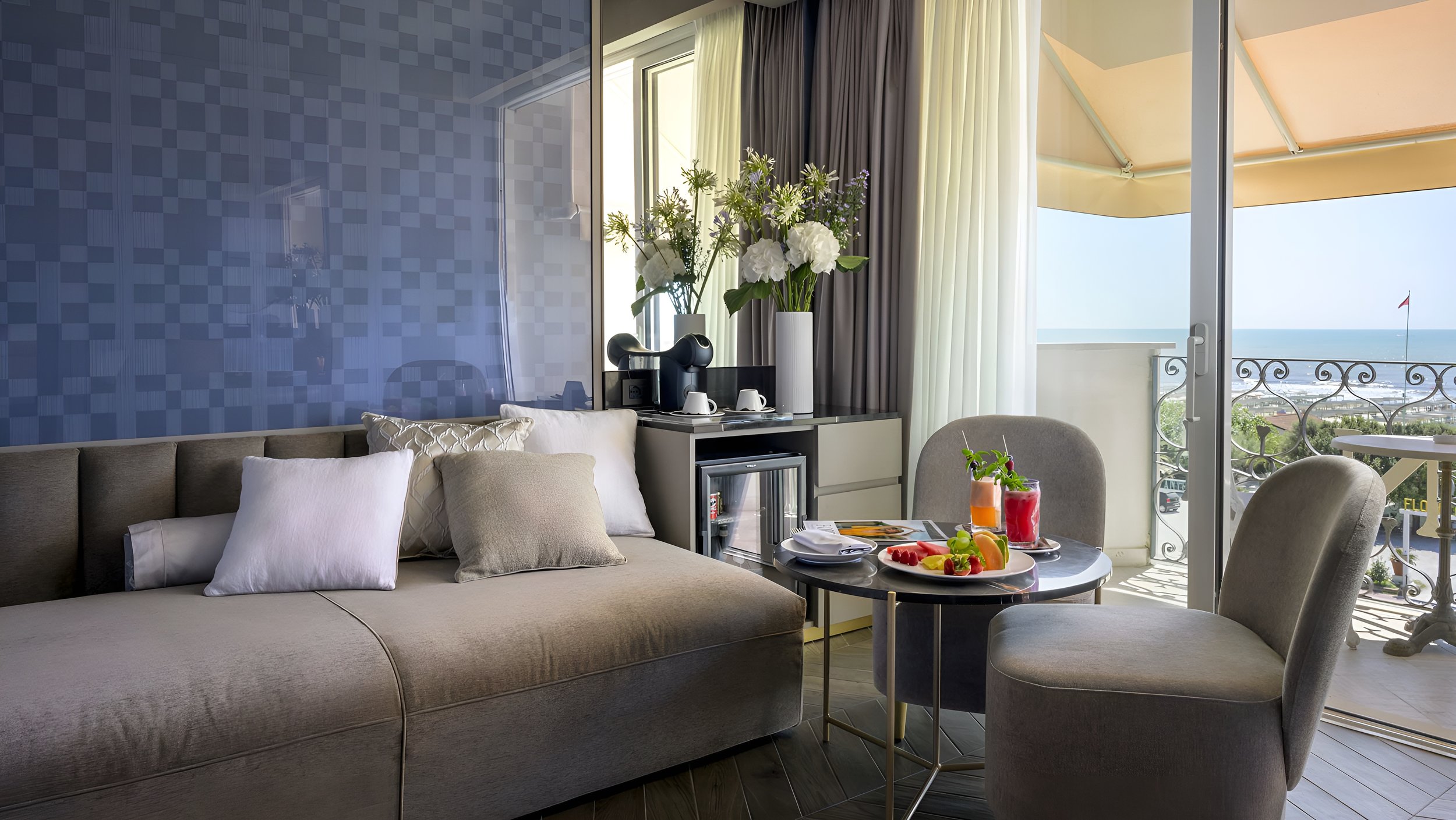 Living room with a beige sofa with white and beige pillows, a small round table with breakfast foods and drinks, a balcony with a view of the ocean, a sliding glass door, and a bright interior with curtains and flowers.