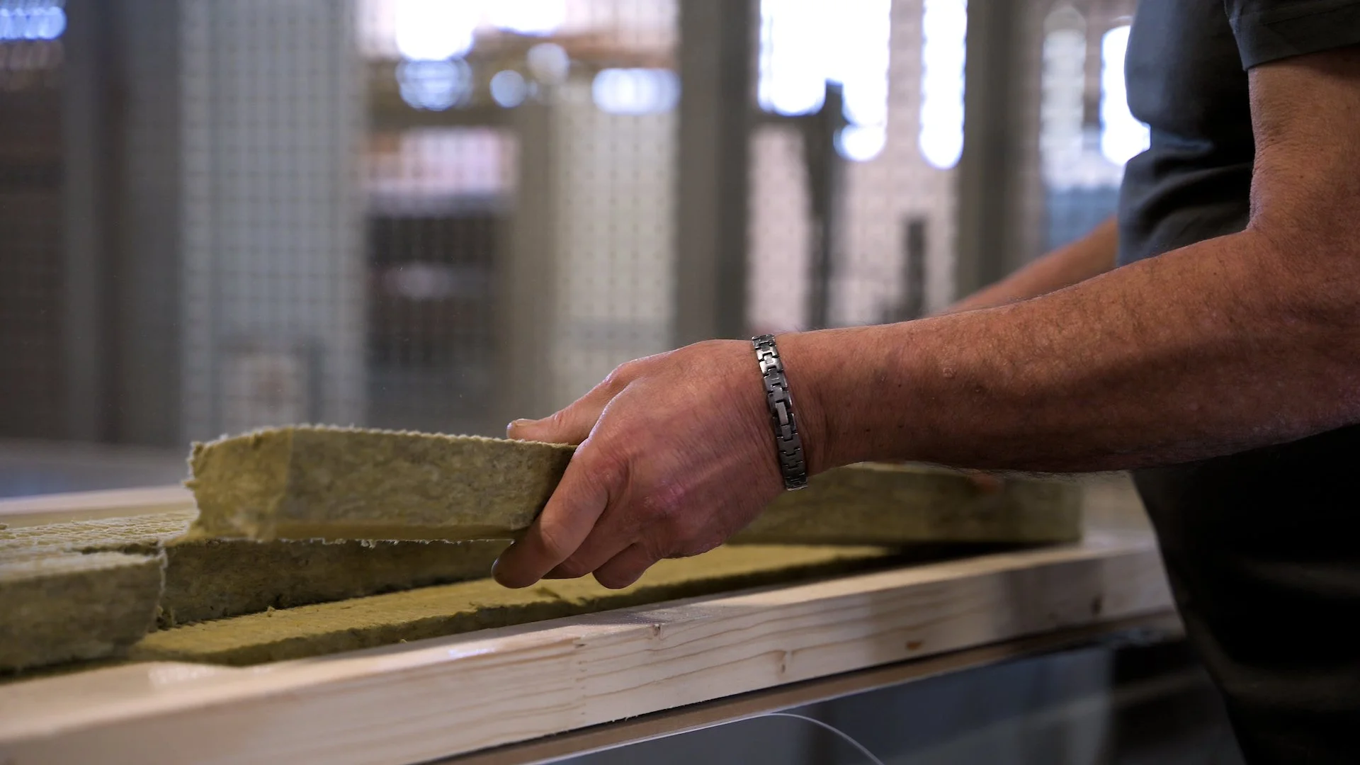A person with a silver bracelet on their left wrist is holding a piece of insulation foam board in a workshop or construction setting. The hand appears to be placed on a wooden surface, and there are metal and wooden structures with blurred lights in the background.