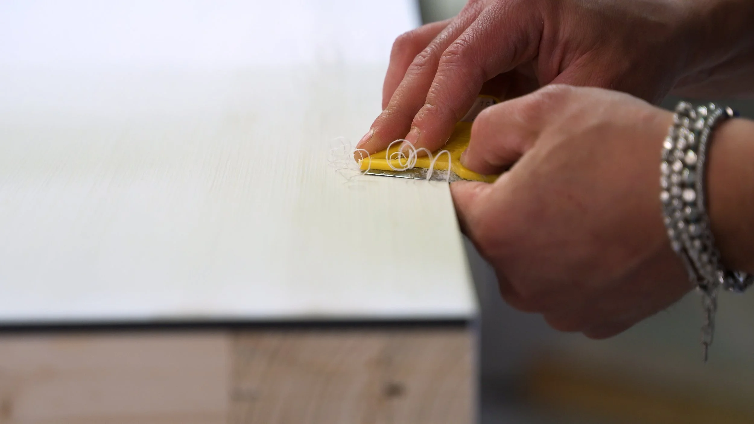 Close-up of a hand using a yellow scraper or tool to work on a surface, with curled threads or fibers on the surface.