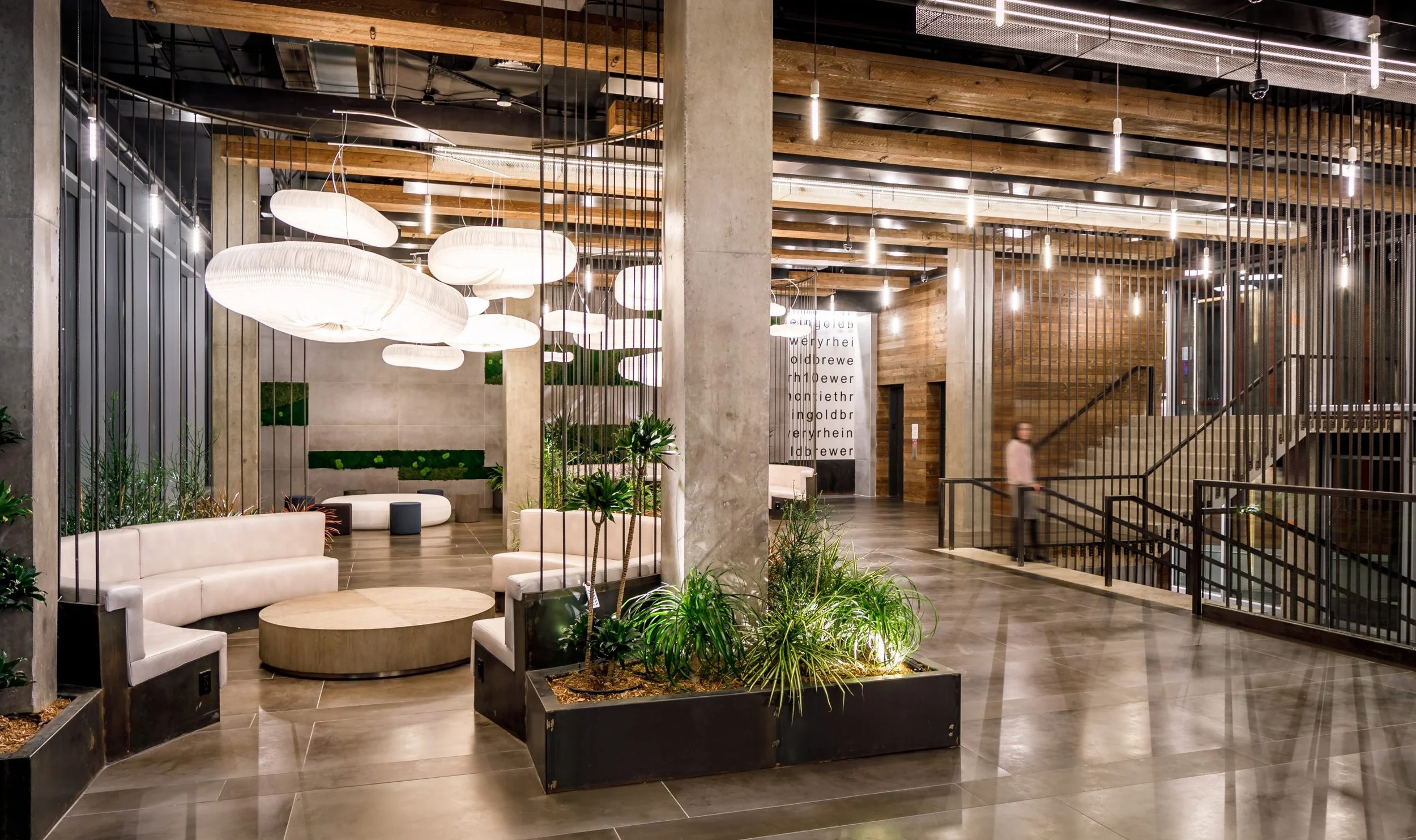 Interior of a modern building lobby with seating areas, potted plants, unique white pendant lights, wooden ceiling beams, metal and concrete structures, and a staircase.