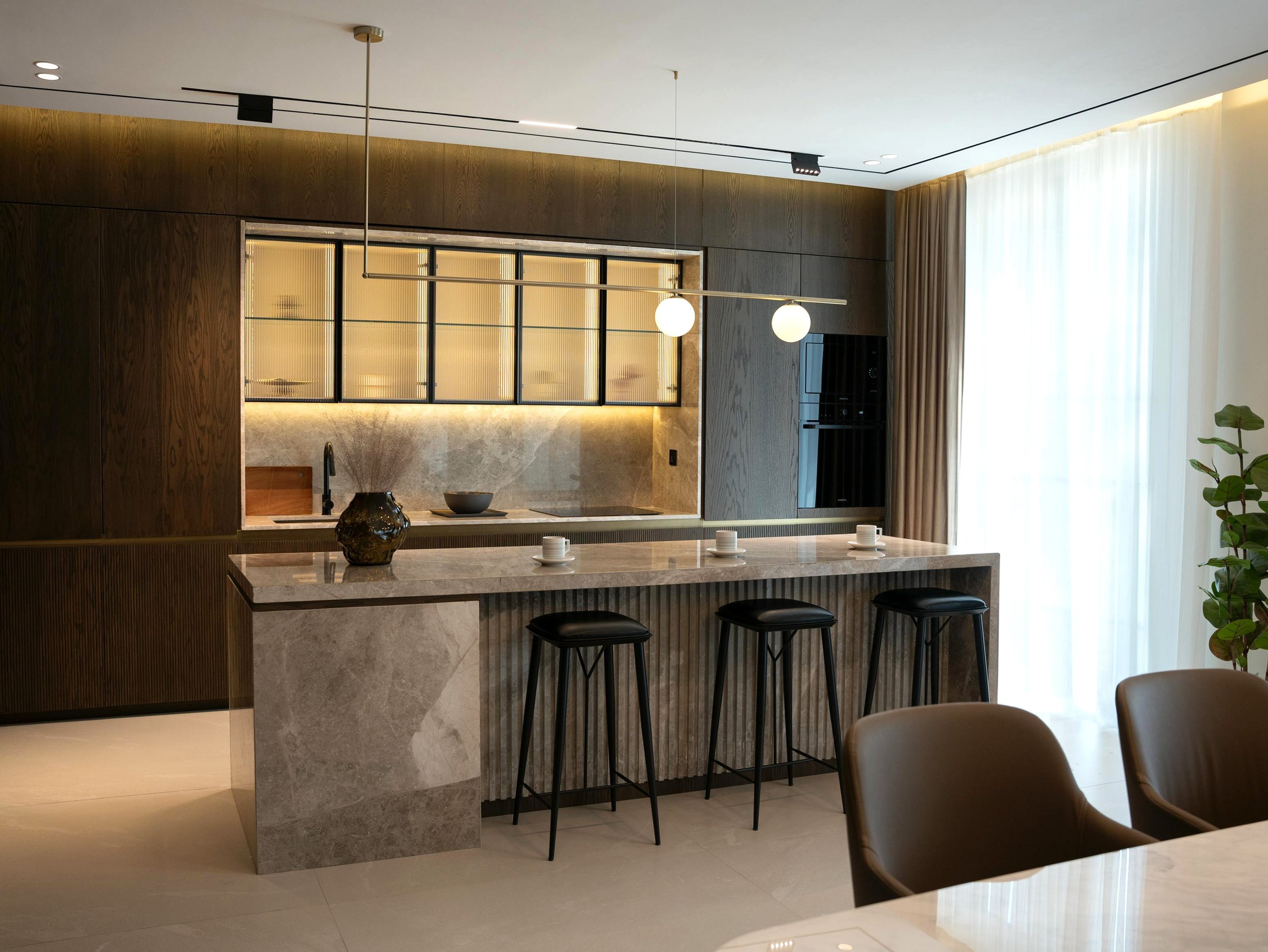 Modern kitchen with a marble island, black stools, wooden cabinetry, and pendant lighting, adjacent to a dining area with large windows and flowing curtains.