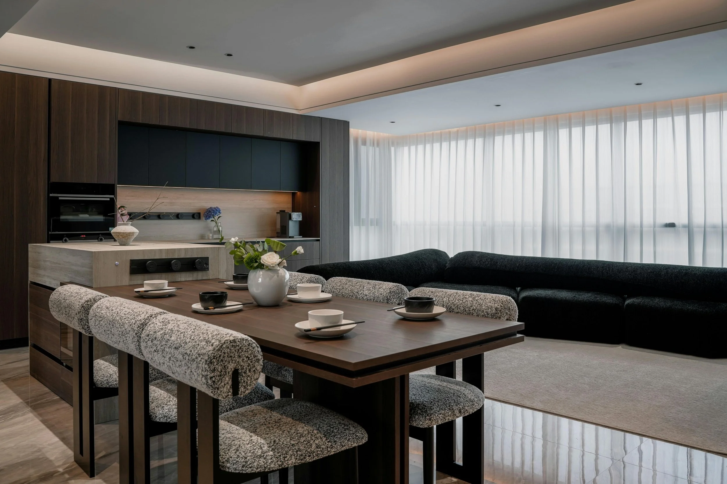 Modern dining and living area with a wooden table set with bowls and cups, a vase with flowers, a large sofa, and a bright window with sheer curtains.