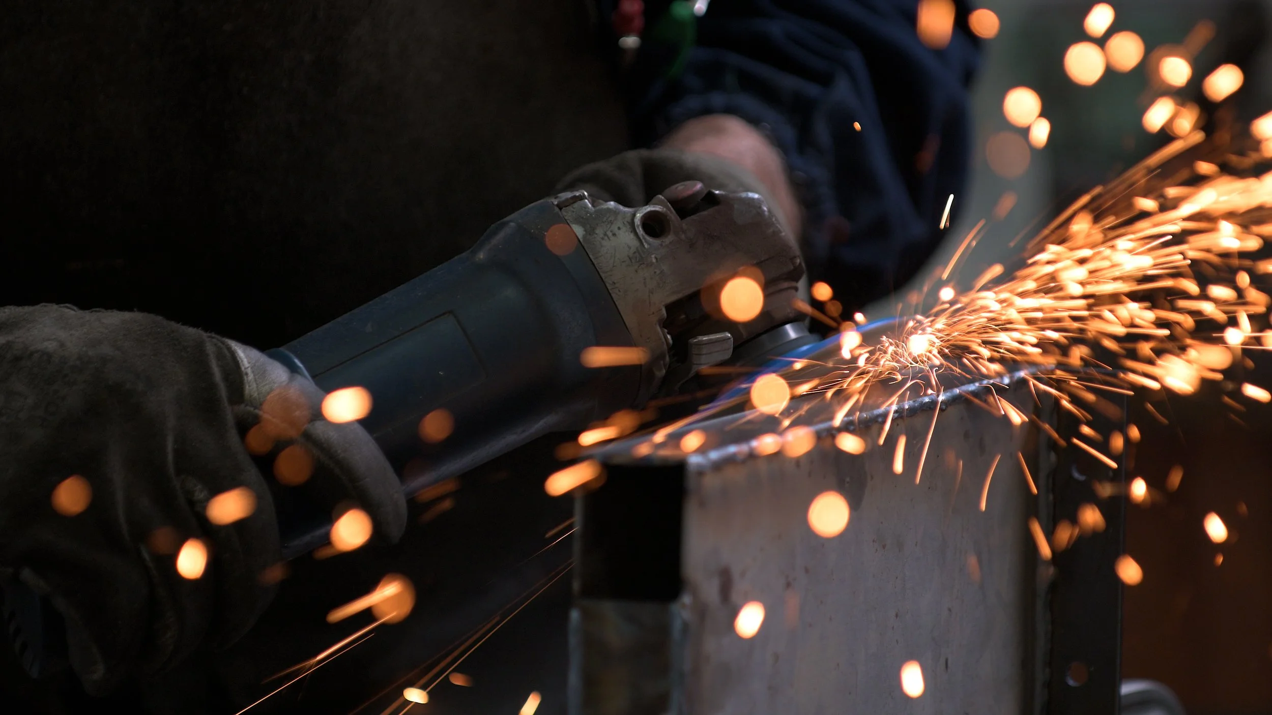 A person wearing gloves uses an angle grinder to cut metal, producing sparks.