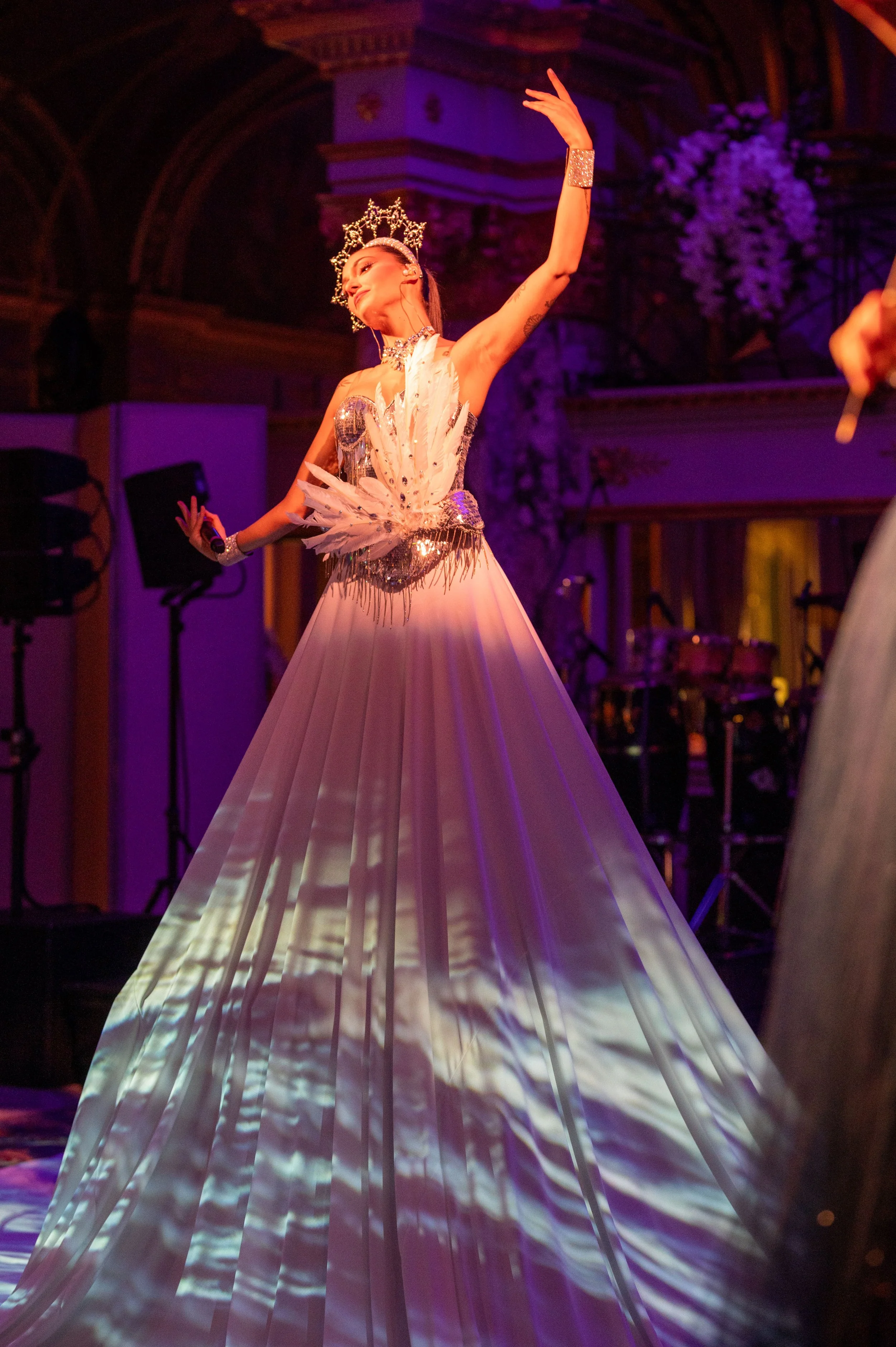 Une femme en tenue de danse portant une couronne brillante effectue une pose artistique dans une ambiance de théâtre ou de spectacle, avec des éclairages colorés et un décor sophistiqué en arrière-plan.