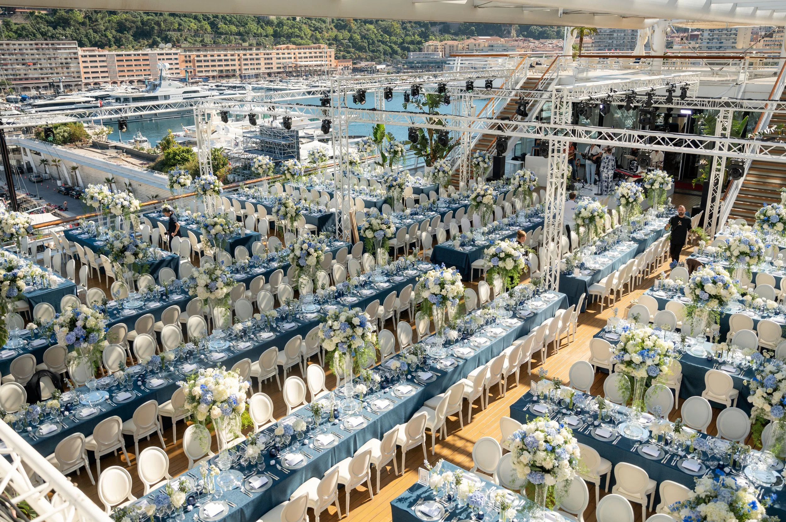 Une grande salle de réception décorée pour un événement avec des tables ornées de fleurs blanches et bleues, sur une terrasse avec vue sur un port rempli de yachts, en arrière-plan des bâtiments résidentiels et une colline verdoyante.