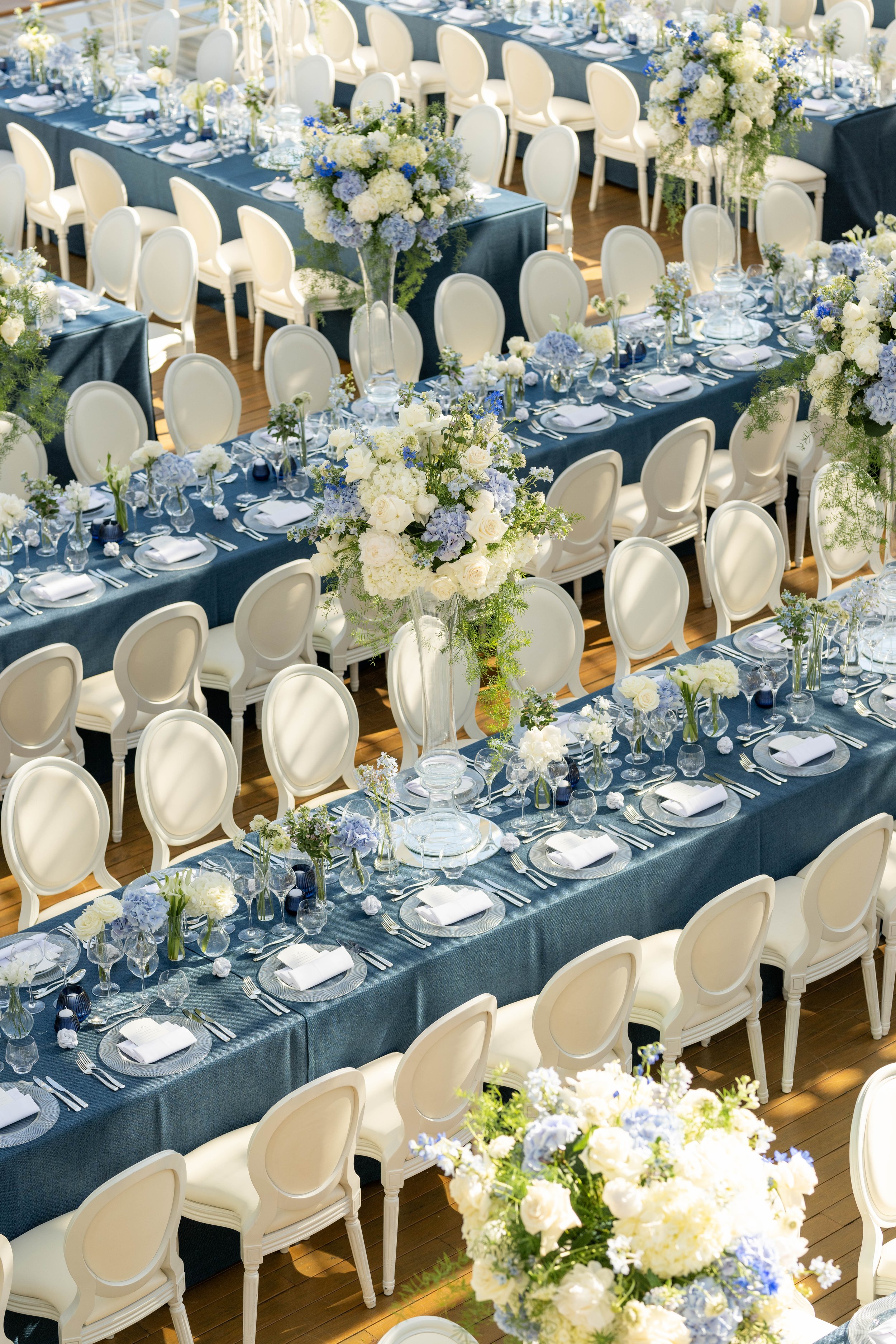 Salle de réception élégante avec longues tables ornées de nappes bleues, décorées de bouquets de fleurs blanches et bleues, entourées de chaises blanches, pour un événement sophistiqué.