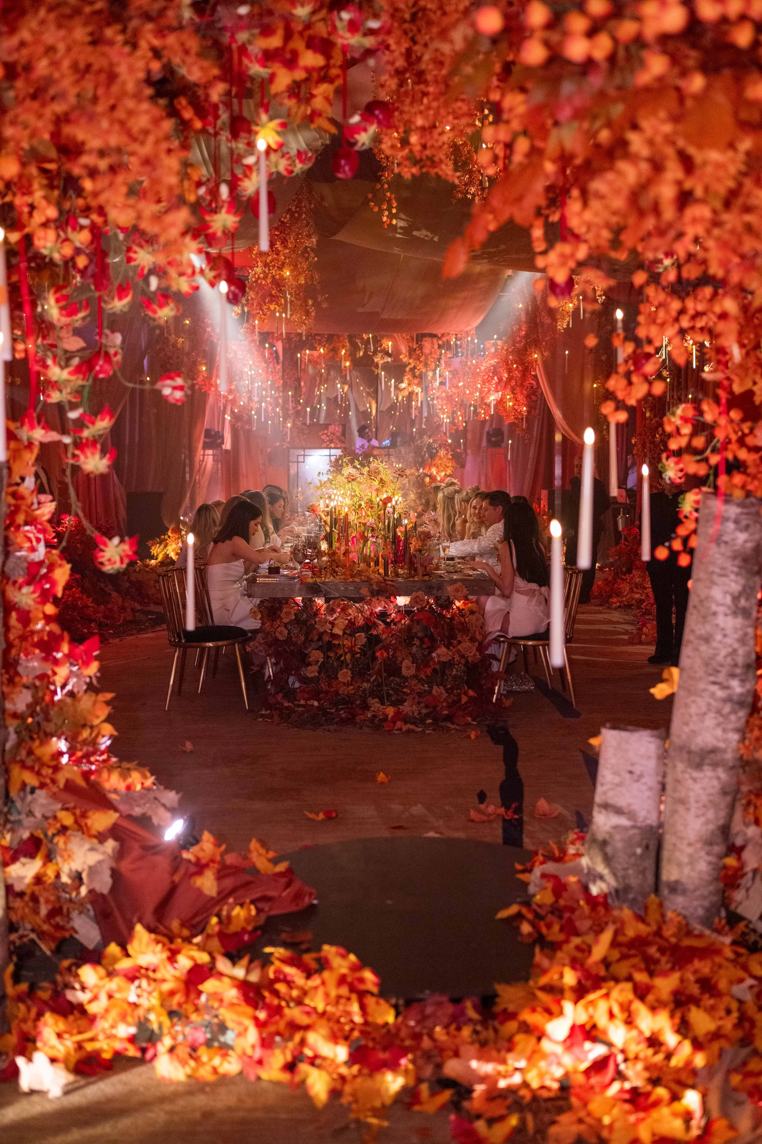 Une salle de banquet richement décorée avec des fleurs orange et rouge suspendues et des bougies, où des personnes sont assises autour d'une longue table pour un dîner élégant.