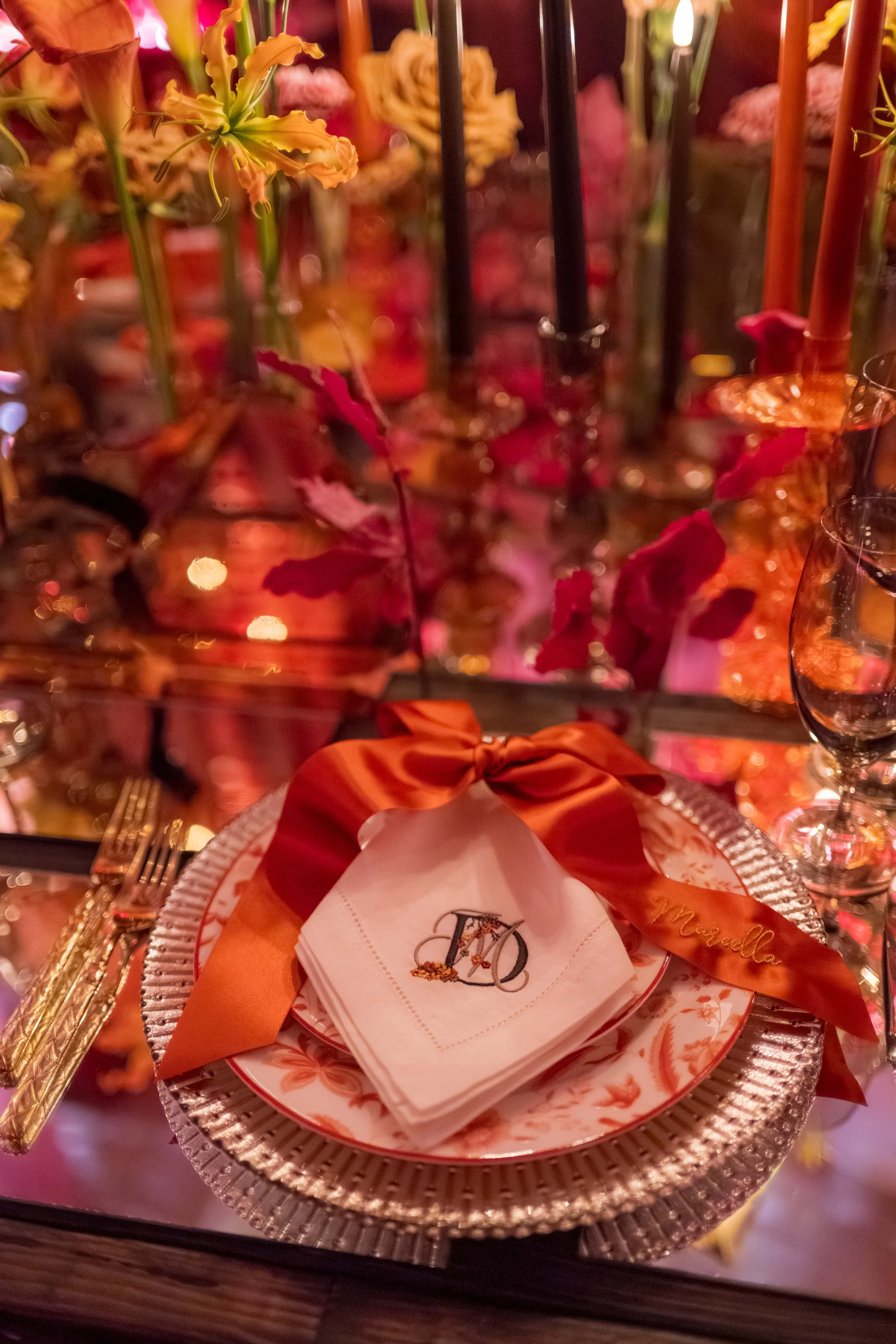 Une table de dîner élégante avec une assiette décorative contenant des napperons en tissu blanc avec une initiale brodée, une serviette roulée avec un ruban d'organdi orange, des couverts en or, une assiette en porcelaine et un verre à vin. En arrièr