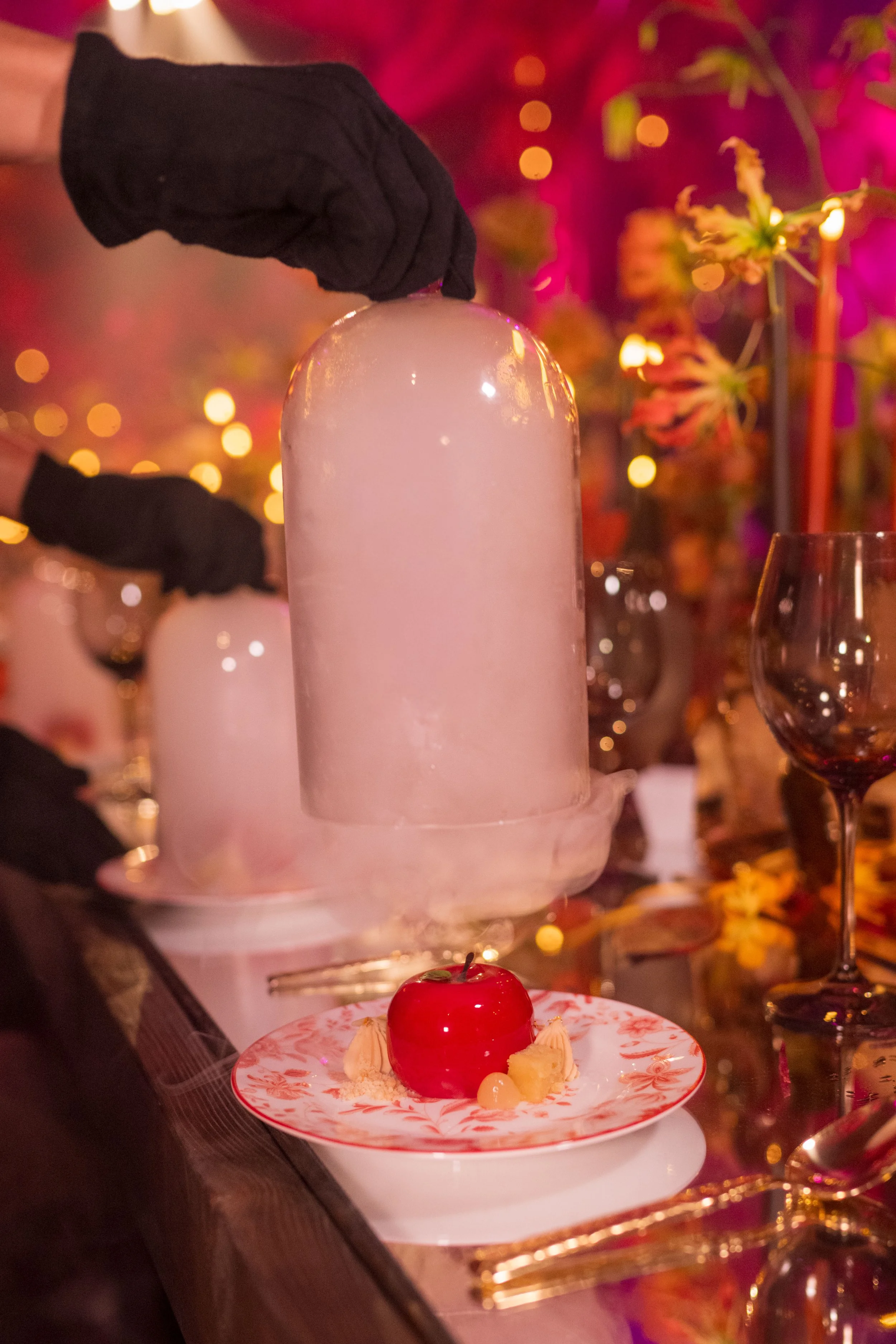 Un gâteau en forme de sphère rouge sur une assiette avec décoration de Noël, entouré de desserts et de verres à vin en arrière-plan dans une ambiance chaleureuse et festive.
