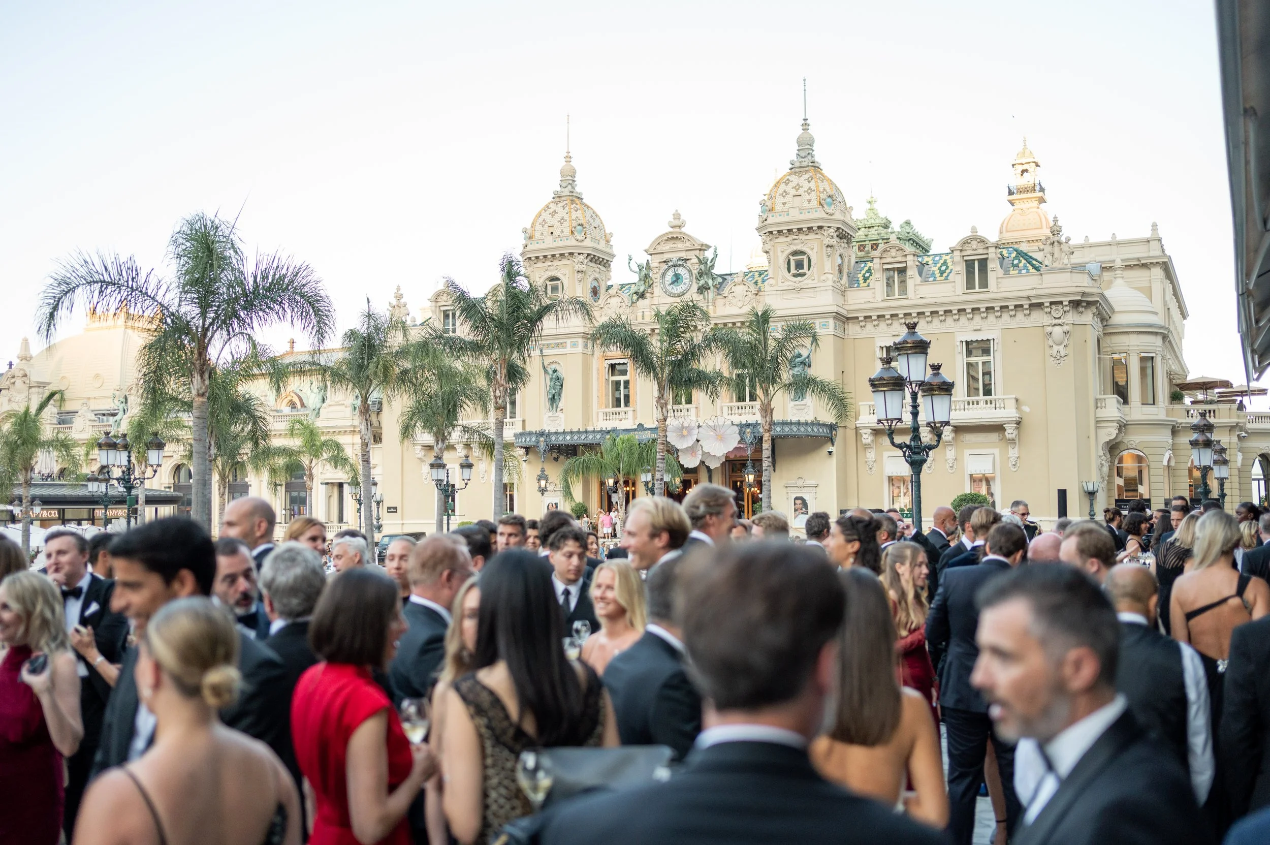 Une foule de personnes habillées en costume de soirée lors d'un événement formel devant un bâtiment historique avec des tourelles et des palmiers.