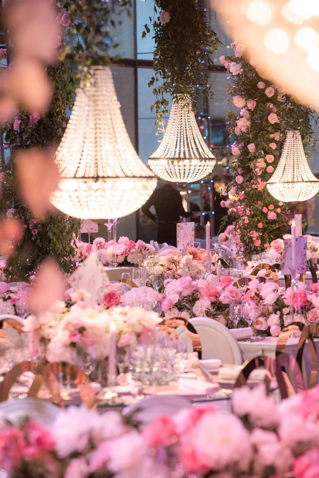 Une salle de mariage élégante décorée de lampes chandeliers en cristal suspendues, et de nombreuses fleurs roses et blanches ornant la salle, avec des tables élégantes prêtes à accueillir les invités.