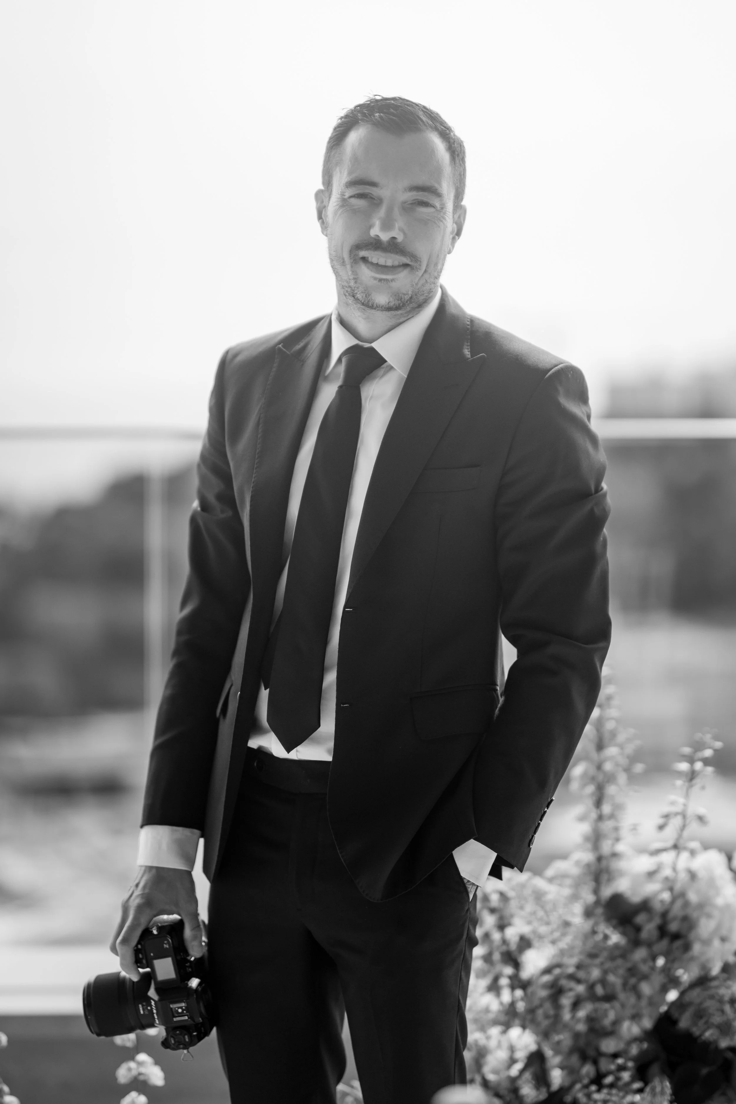 Homme en costume noir, chemise blanche et cravate, tenant un appareil photo, souriant, en plein air, arrière-plan flou avec des plantes et un bâtiment en arrière-plan.