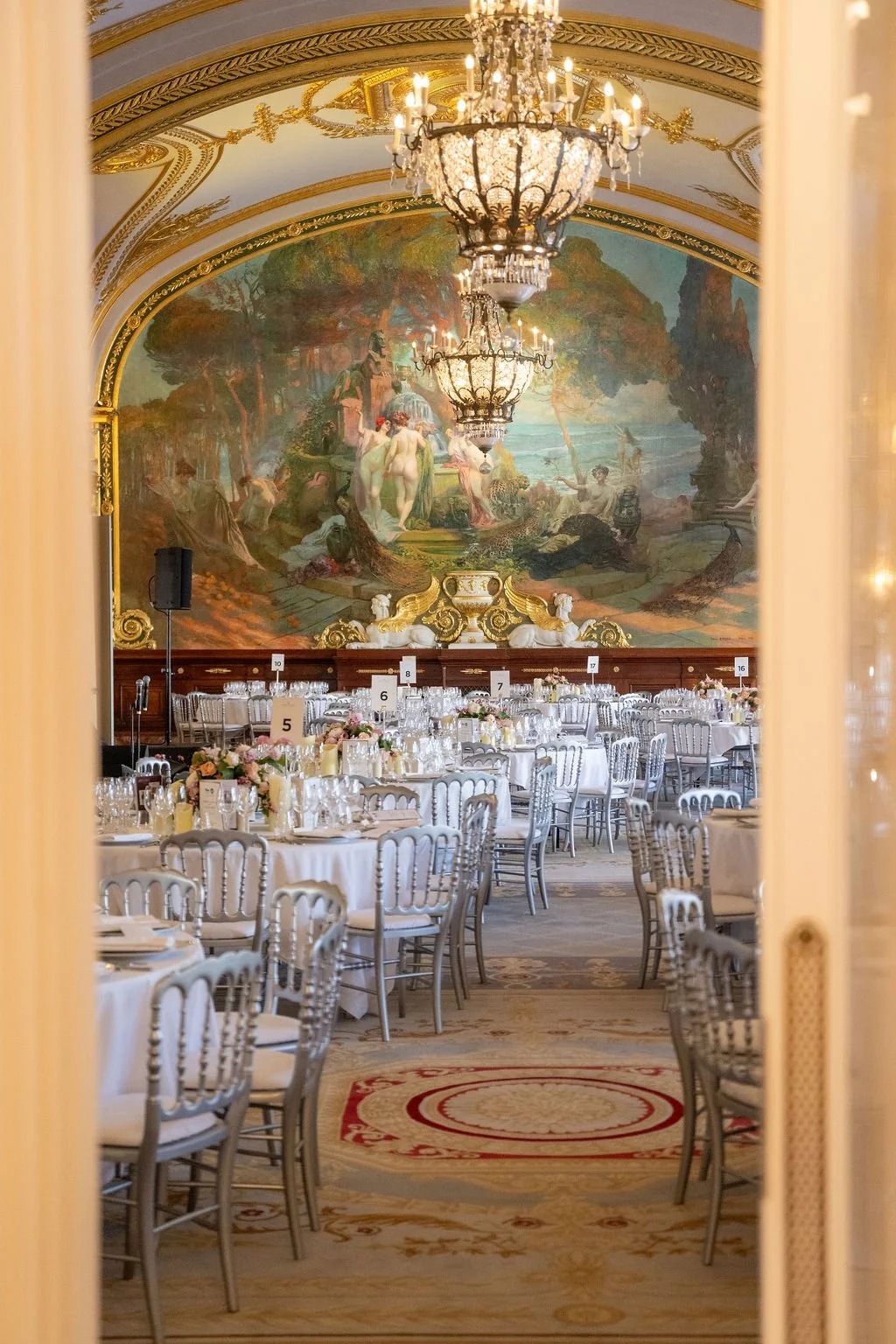 Salle de banquet élégante avec des tables rondes habillées de nappes blanches, décorées avec des fleurs et des bougies, sous un grand plafond orné de fresques classiques et éclairée par deux grands lustres en cristal.