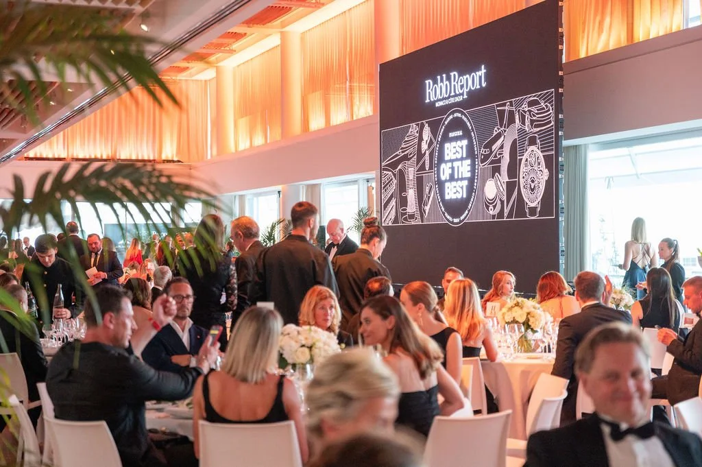 Gens assis et debout, déjeunant ou réseautant lors d'un événement formel dans une salle élégante, avec un grand écran noir affichant le logo 'Robb Report' et le texte 'Best of the Best', décorée avec des fleurs blanches sur chaque table.