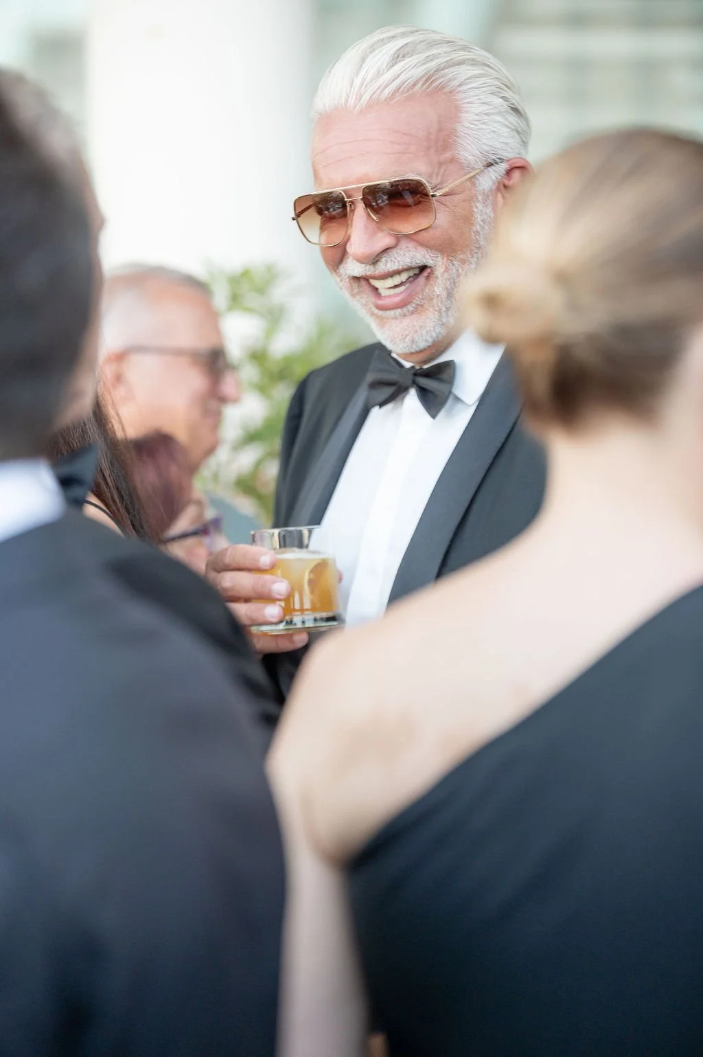 Un homme âgé en smoking, portant des lunettes de soleil, souriant et tenant un verre de cocktail, en compagnie de plusieurs autres personnes lors d'une fête élégante.