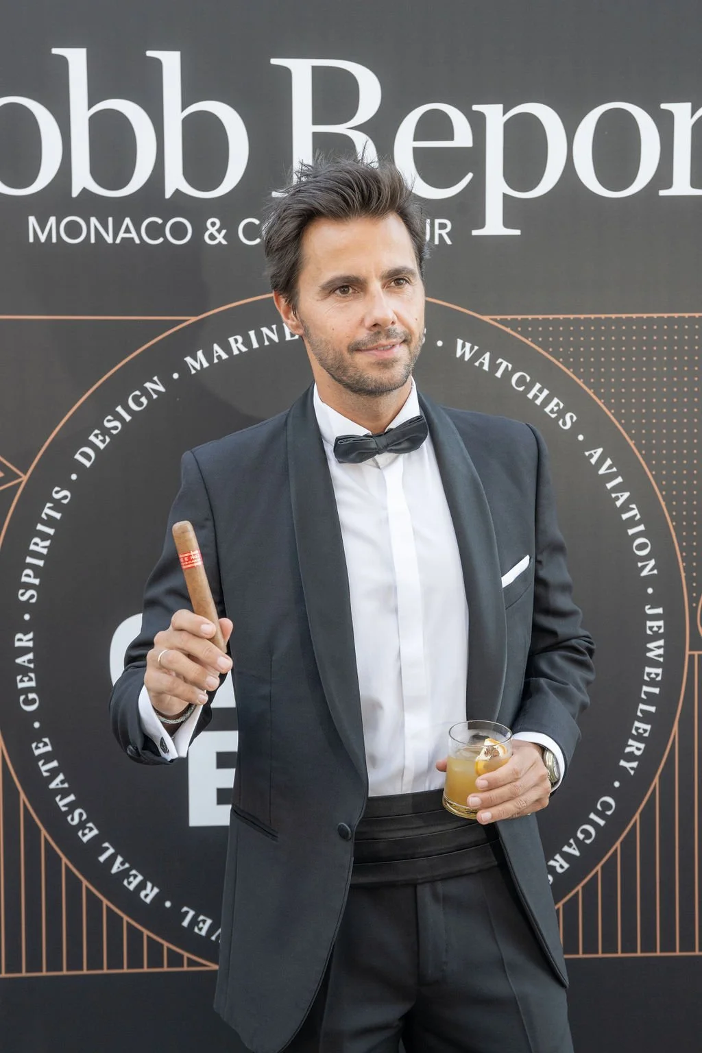 Homme en smoking noir avec nœud papillon, tenant un cigare dans une main et un verre de cocktail dans l'autre, posant devant un panneau noir avec des inscriptions promotionnelles.