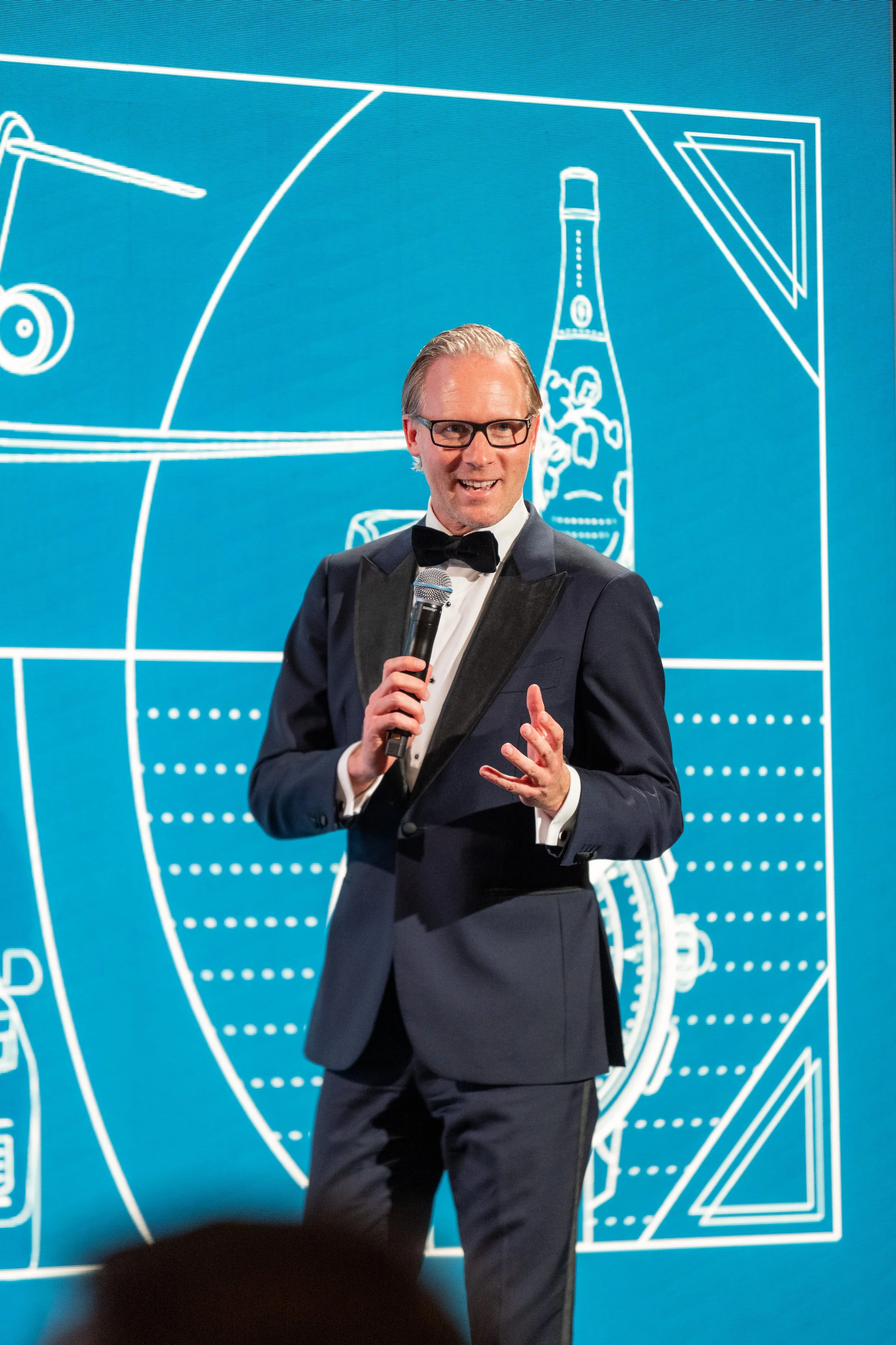 Homme en smoking avec micro, parlant lors d'une présentation ou d'un événement VIP, fond bleu avec dessins techniques ou schémas.