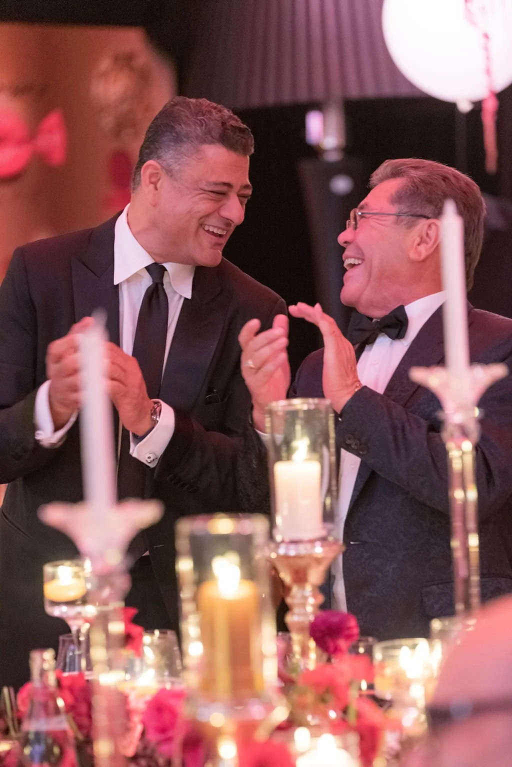 Deux hommes en costume noir et cravate noire, souriants et riant, discutent lors d'une soirée élégante avec des bougies et des décorations florales sur la table.