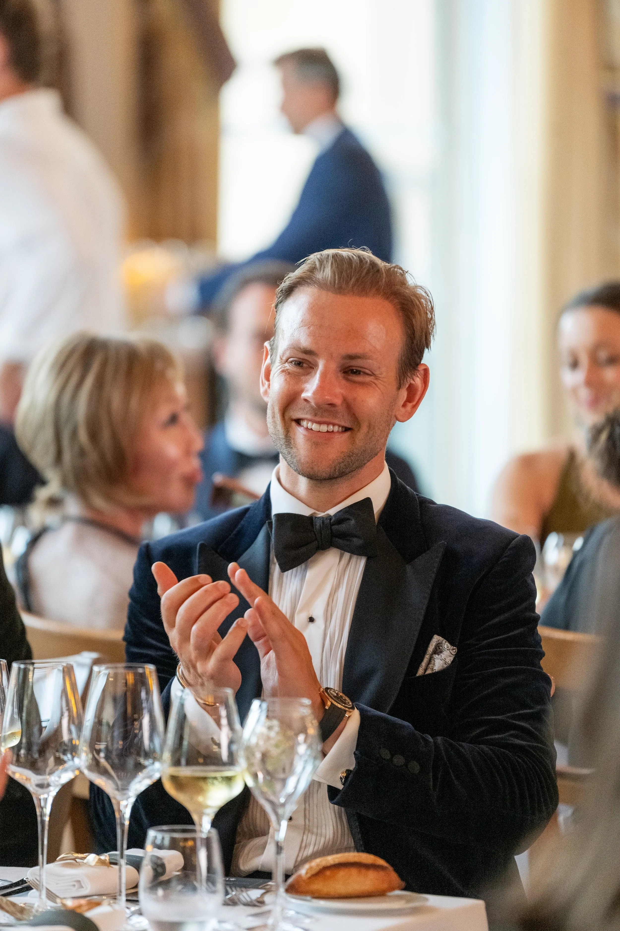 Un homme souriant en smoking noir, assis à une table de restaurant élégante, entouré d'autres convives, avec des verres de vin blanc et du pain sur la table.