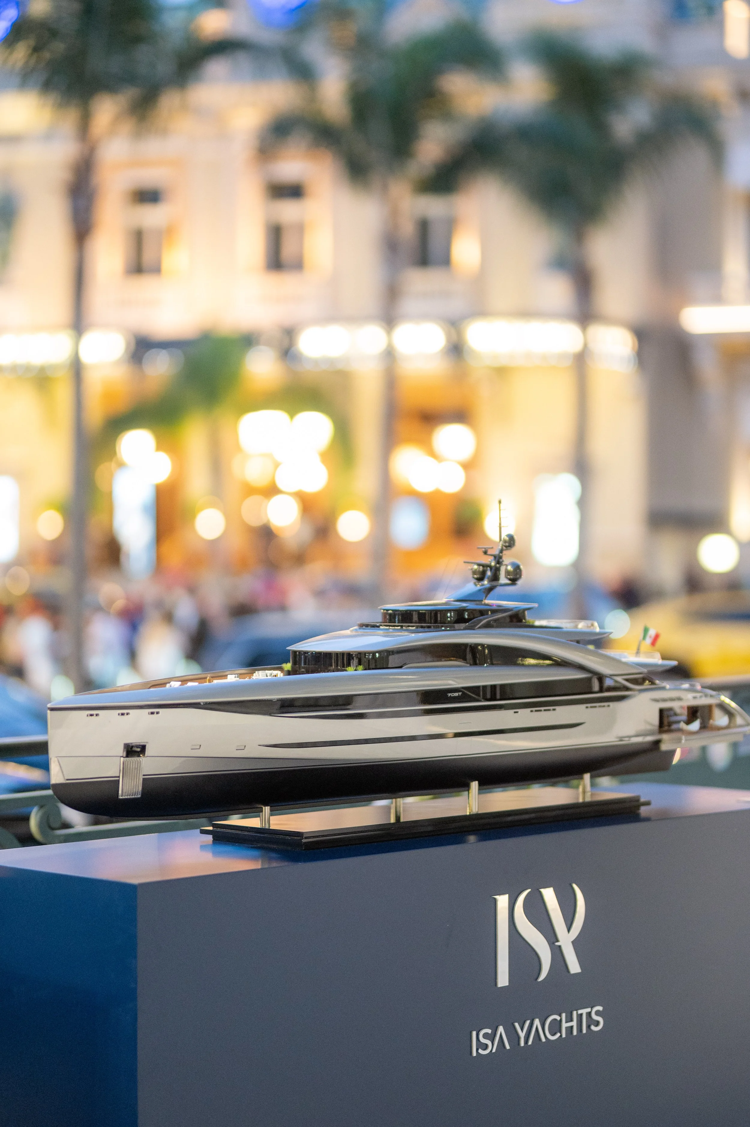 Modèle réduit d'un yacht de luxe noir et gris présenté sur un socle à une exposition, avec un fond flou de bâtiments et d'arbres en ville
