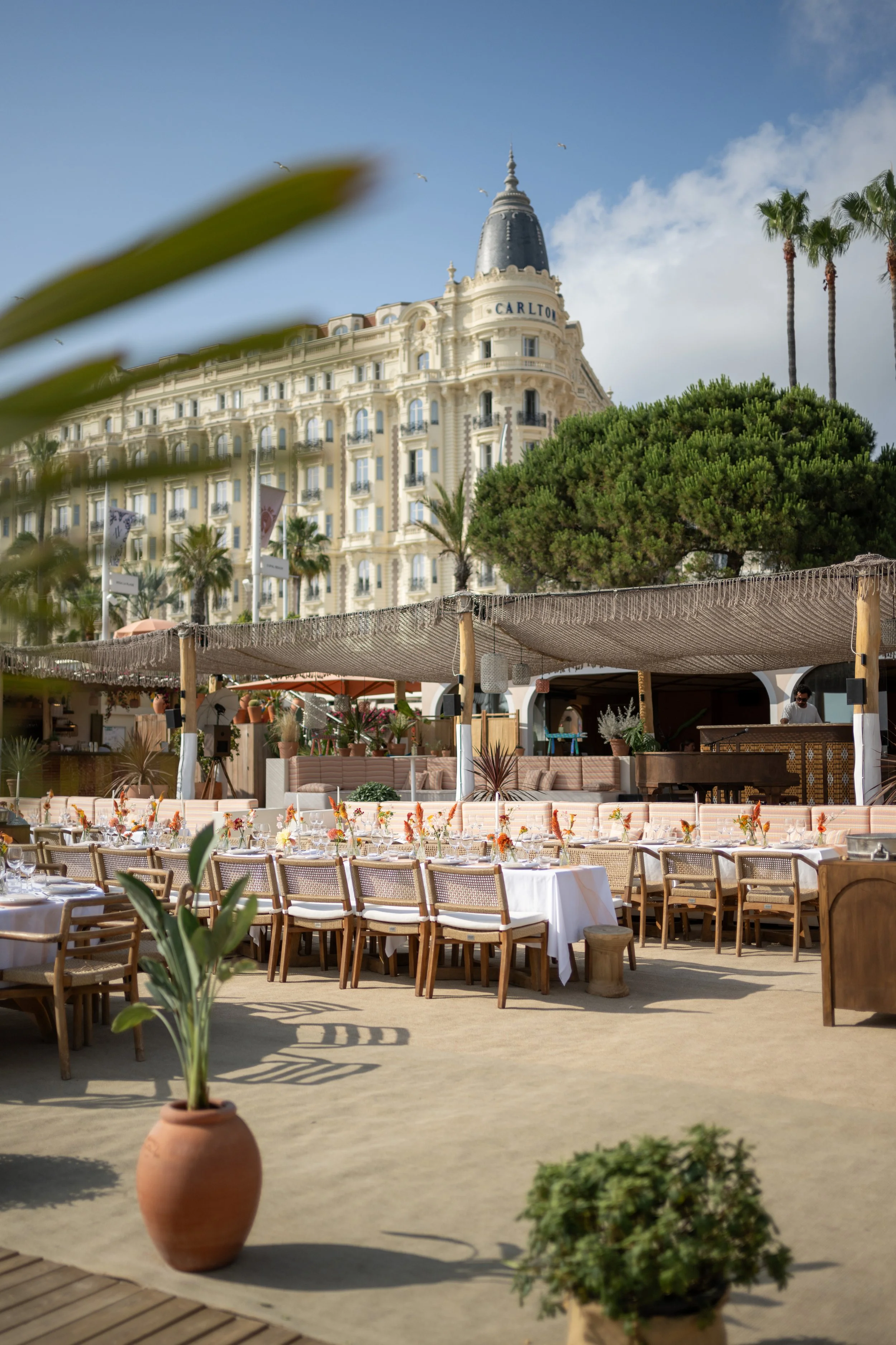 Une terrasse de restaurant en plein air avec des tables ornées de nappes blanches et de fleurs, entourée de pots de plantes, avec en arrière-plan un bâtiment de style hôtelier avec le nom 'Carlton' et des palmiers, sous un ciel partiellement nuageux.
