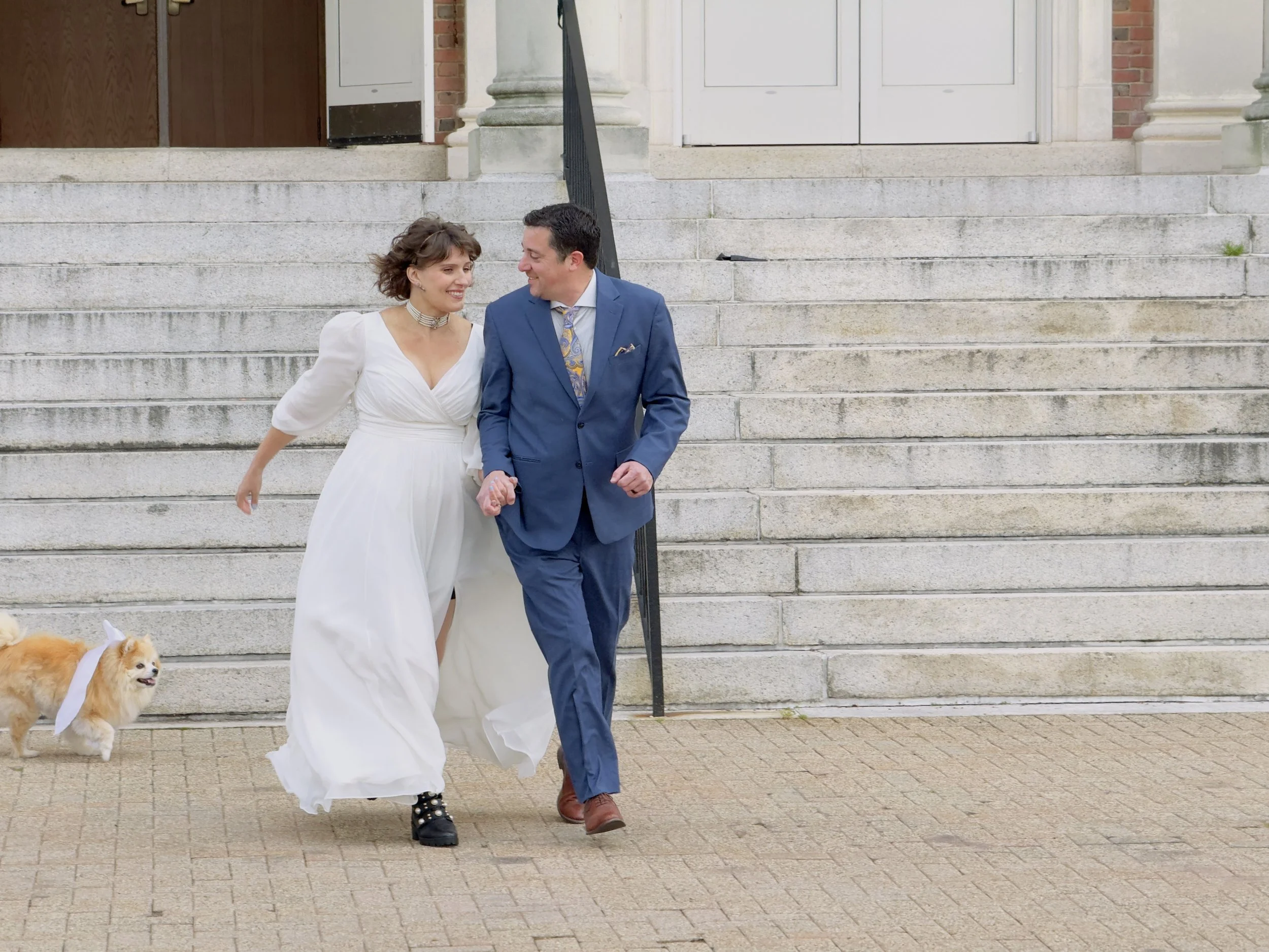 Joyful couple holding hands and walking their dog past stone steps during an outdoor portrait session in Avon, Connecticut.
