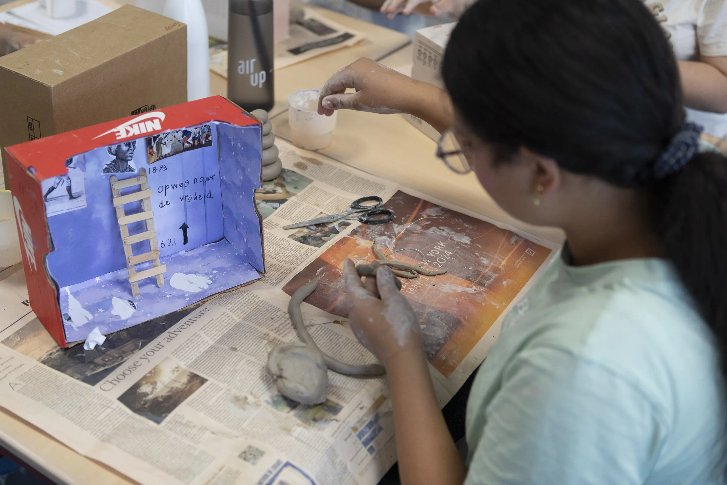 A girl with dark hair and glasses creating a miniature winter scene inside a decorated shoe box, with art supplies and scissors on the table covered in newspaper.