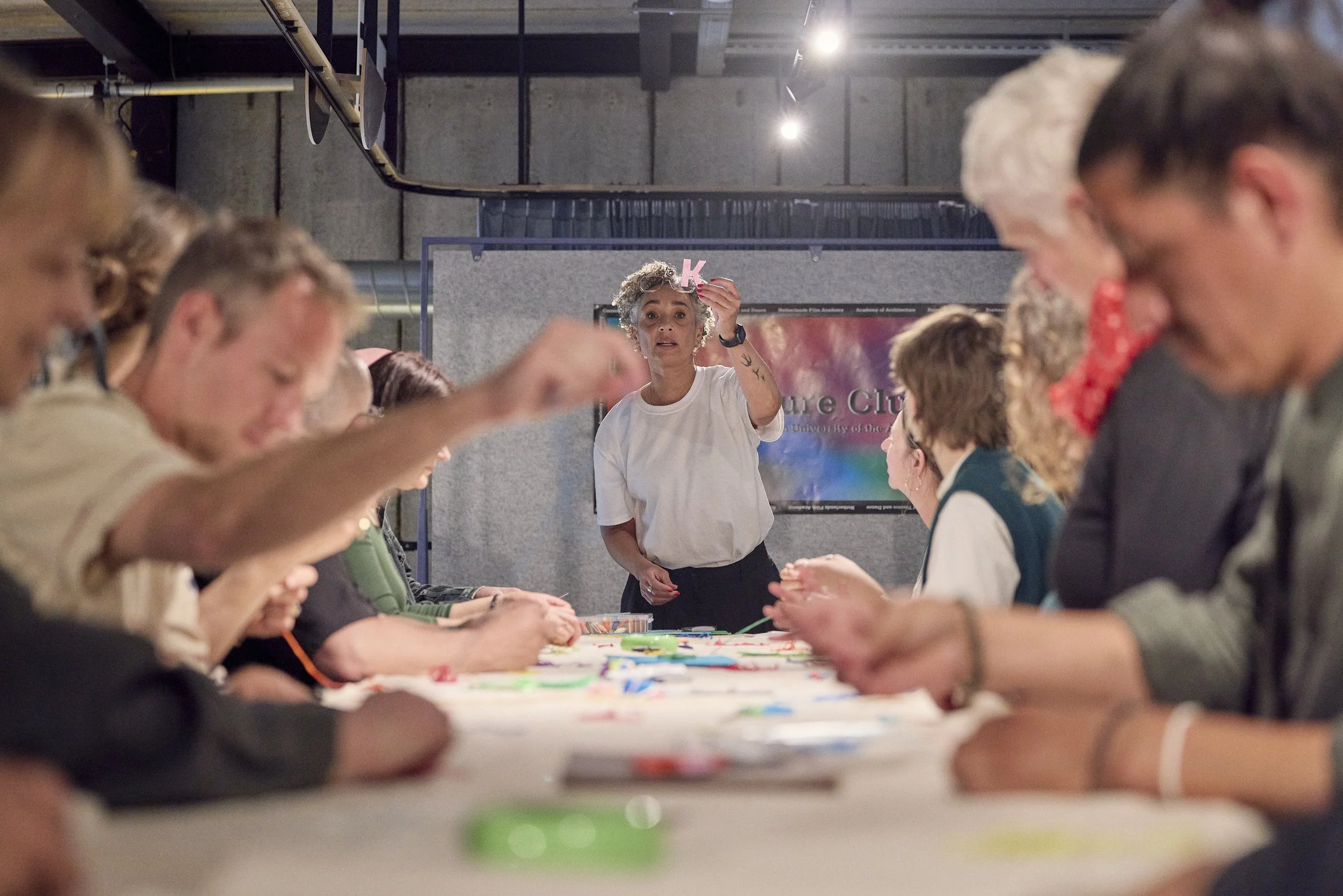 A woman with gray curly hair and tattoos on her arms standing behind a long table, holding a pink letter 'K' prop, surrounded by a group of people sitting at the table in a room with concrete walls and visible ceiling beams.