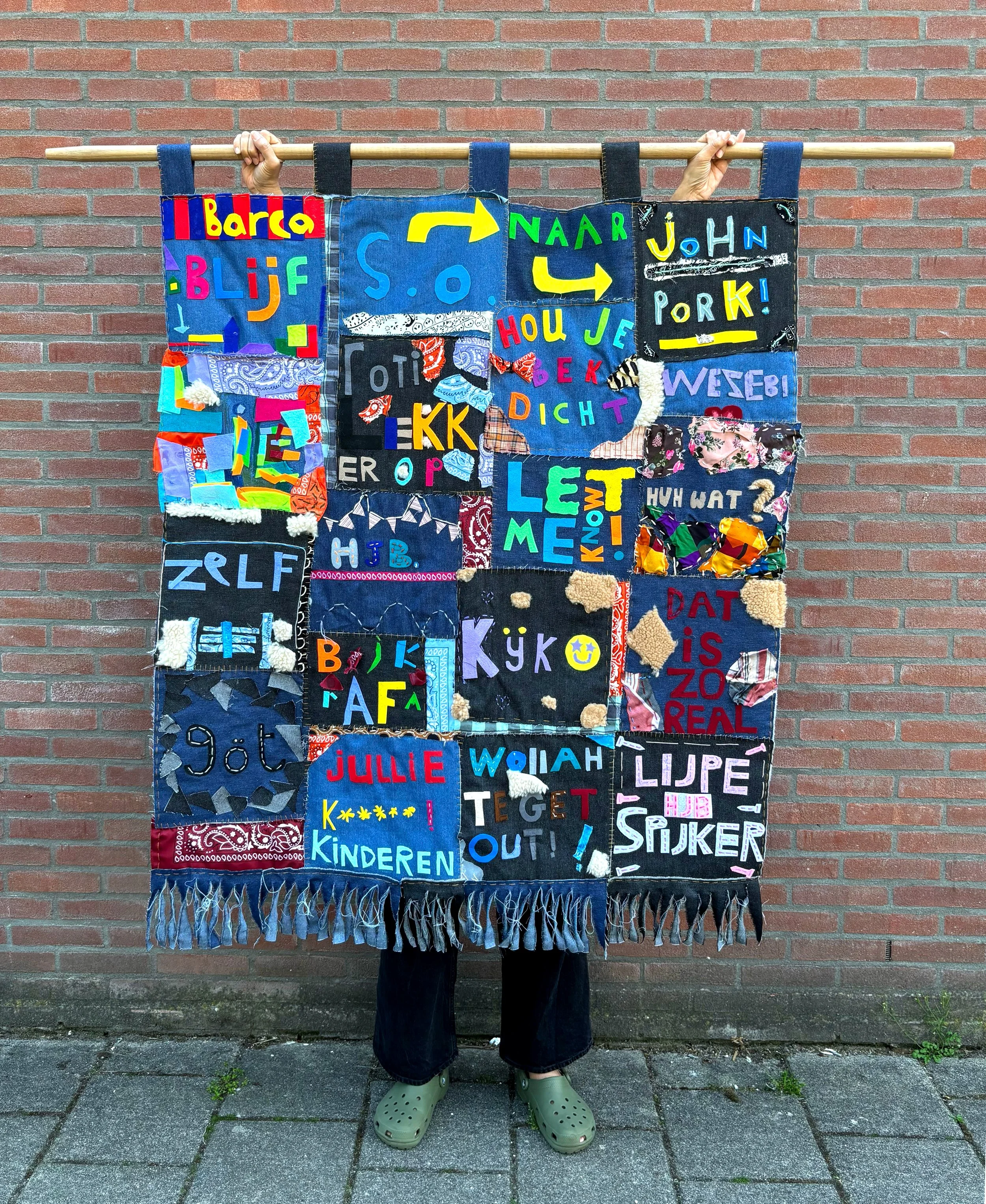 Person holding a colorful fabric art piece with letters and symbols, dressed in dark jeans and green Crocs, standing against a brick wall.