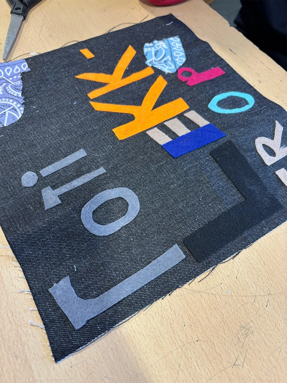 A fabric piece with colorful felt letters and shapes sewn onto a black background, partially spelling 'ROTI LEKKER OP'.