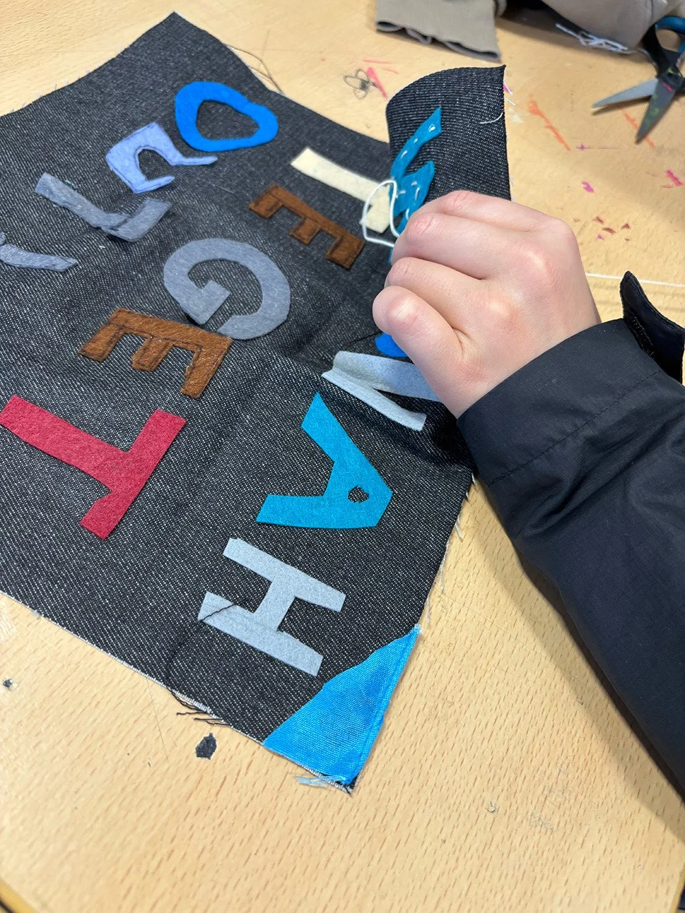 A person sewing felt letters onto a piece of fabric, spelling 'WOLLAH TE GET OUT', on a wooden table.