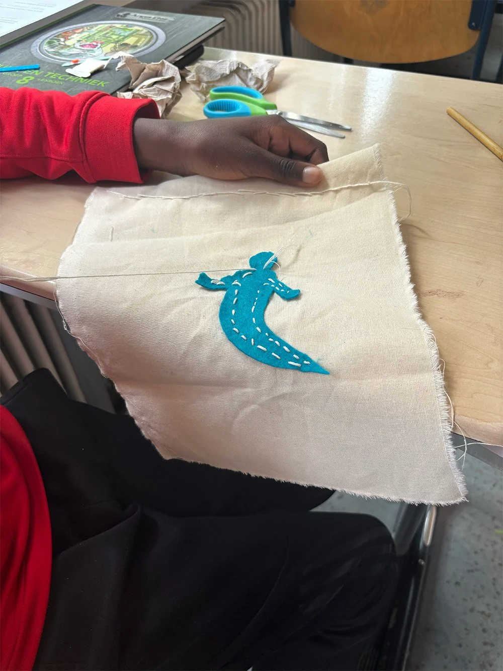 Child sewing a blue fabric dinosaur applique onto a beige cloth piece.