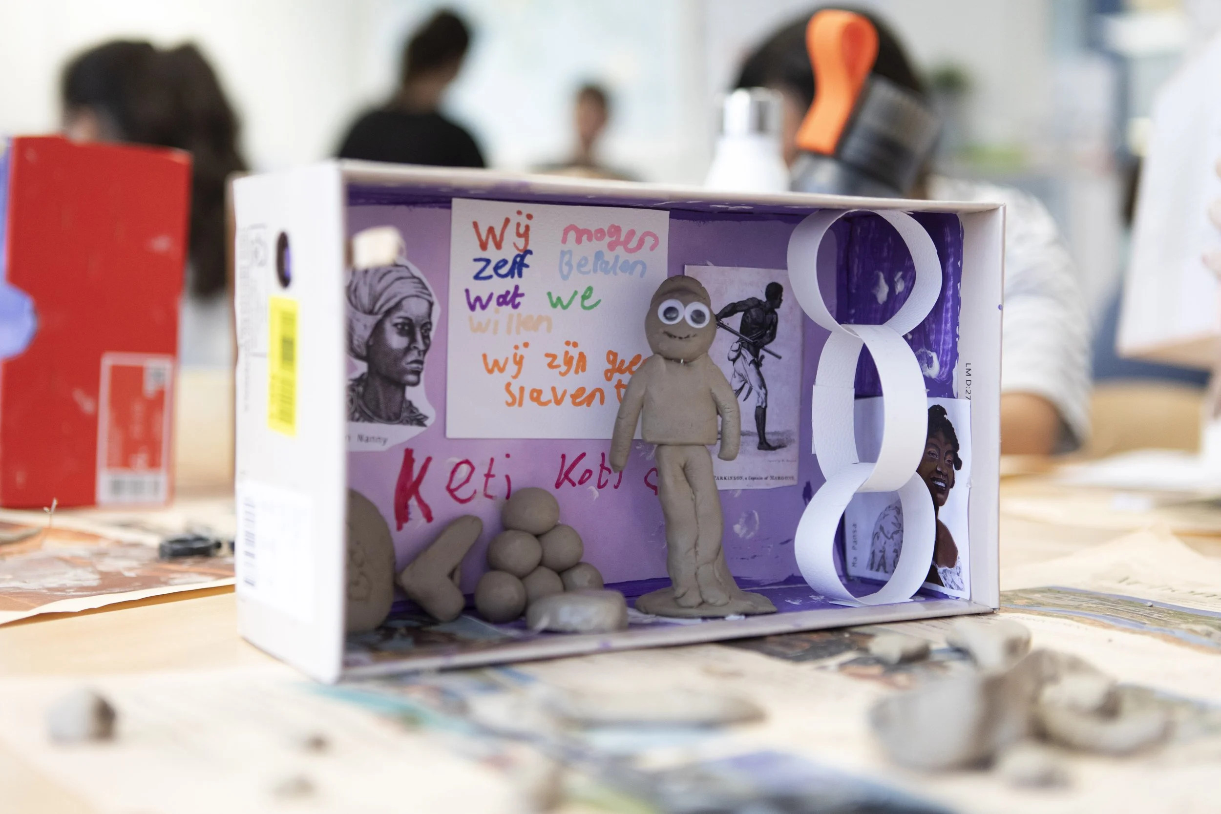 A classroom project - diorama - display featuring clay figures, handwritten notes, and posters with images of women, including one with a headscarf and another smiling woman. Blurred students are in the background.