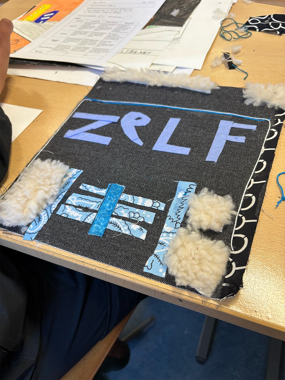 A black fabric display with the word 'ZELF' in purple letters, surrounded by pieces of blue patterned fabric and fluffy white material, on a wooden table with papers, books, and sewing supplies.