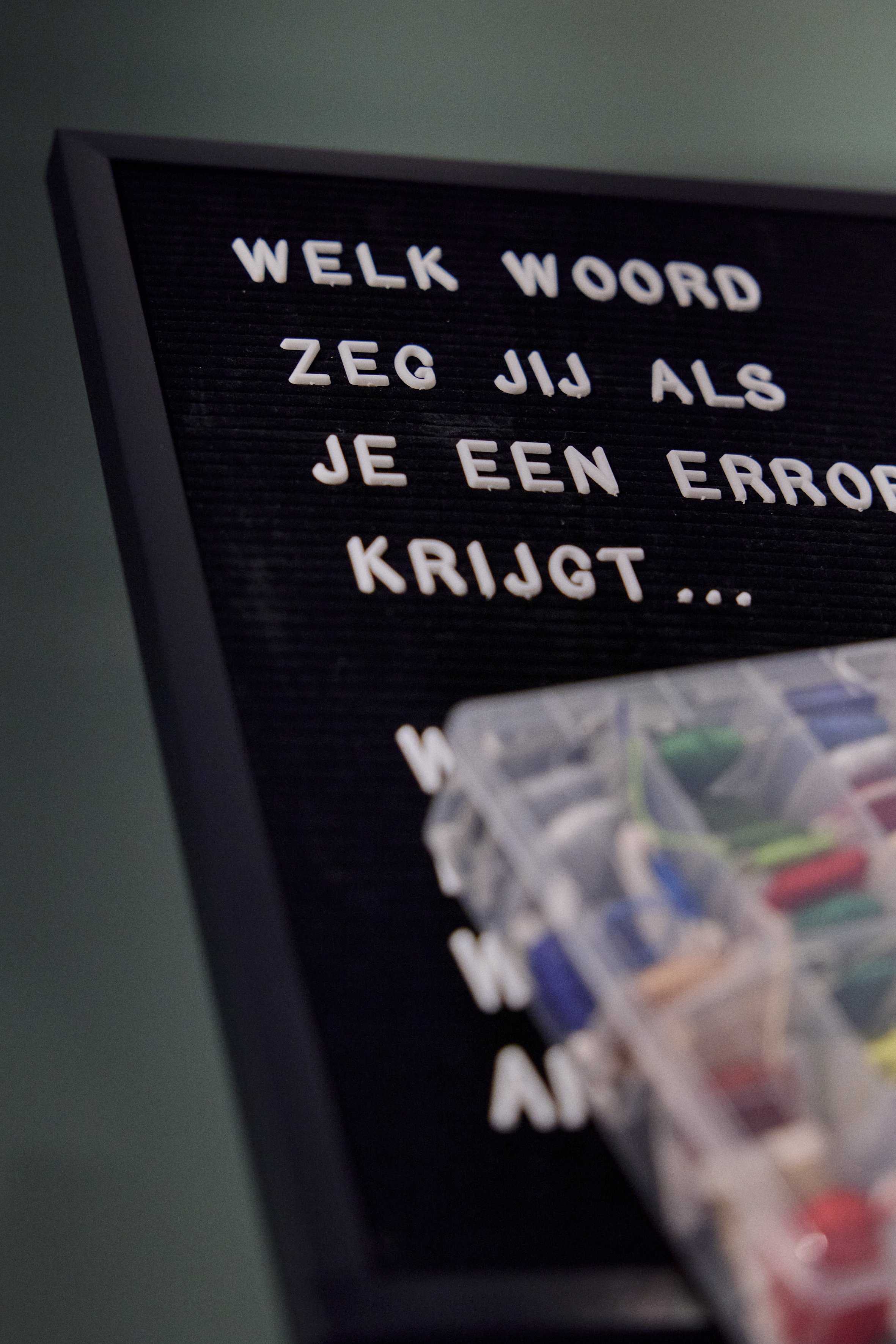 Close-up of a letter board with white magnetic letters spelling out "WELK WOORD ZEG JIJ ALS JE EEN ERROR KRIJGT" and part of a colorful puzzle piece in the lower right corner.