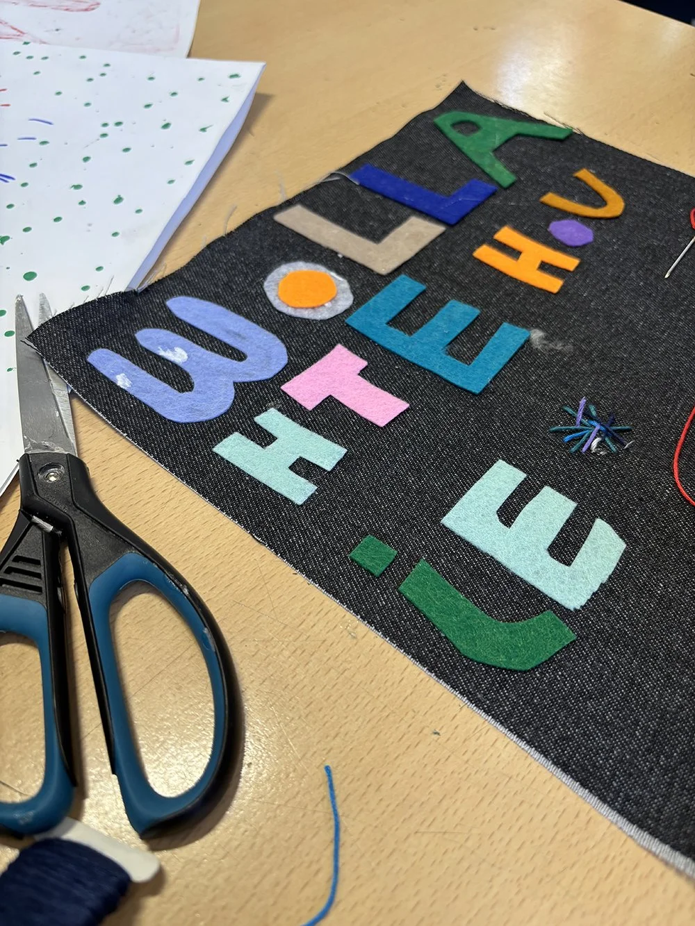 Colorful felt letters spelling ''WOLLAH TE GET OUT''. Scissors, pins, and fabric on a wooden table.