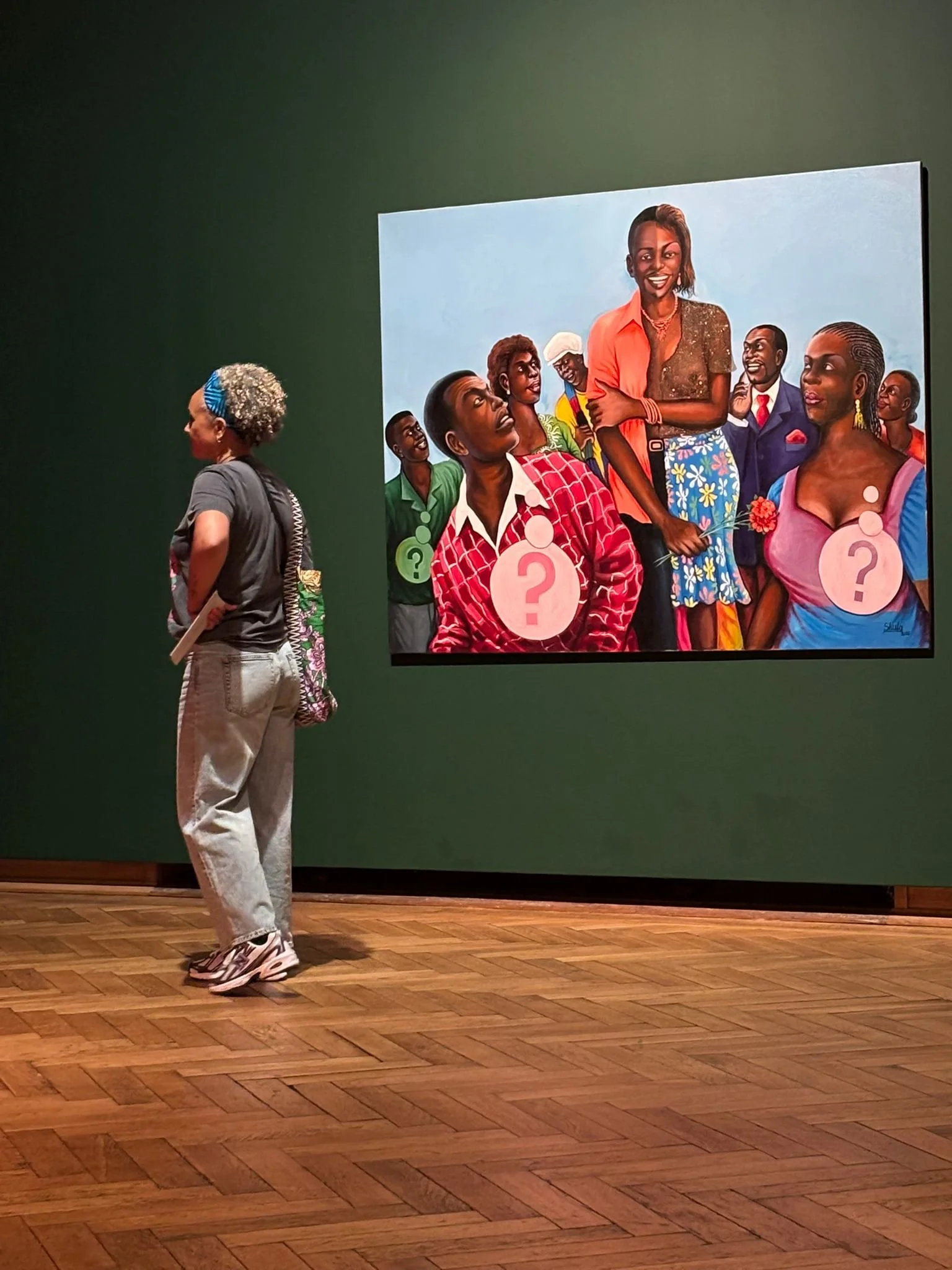 A woman with short curly gray hair wearing a blue headband, gray T-shirt, and beige pants is standing and looking at a large colorful painting in an art gallery. The painting depicts diverse people with question mark symbols on their chests.