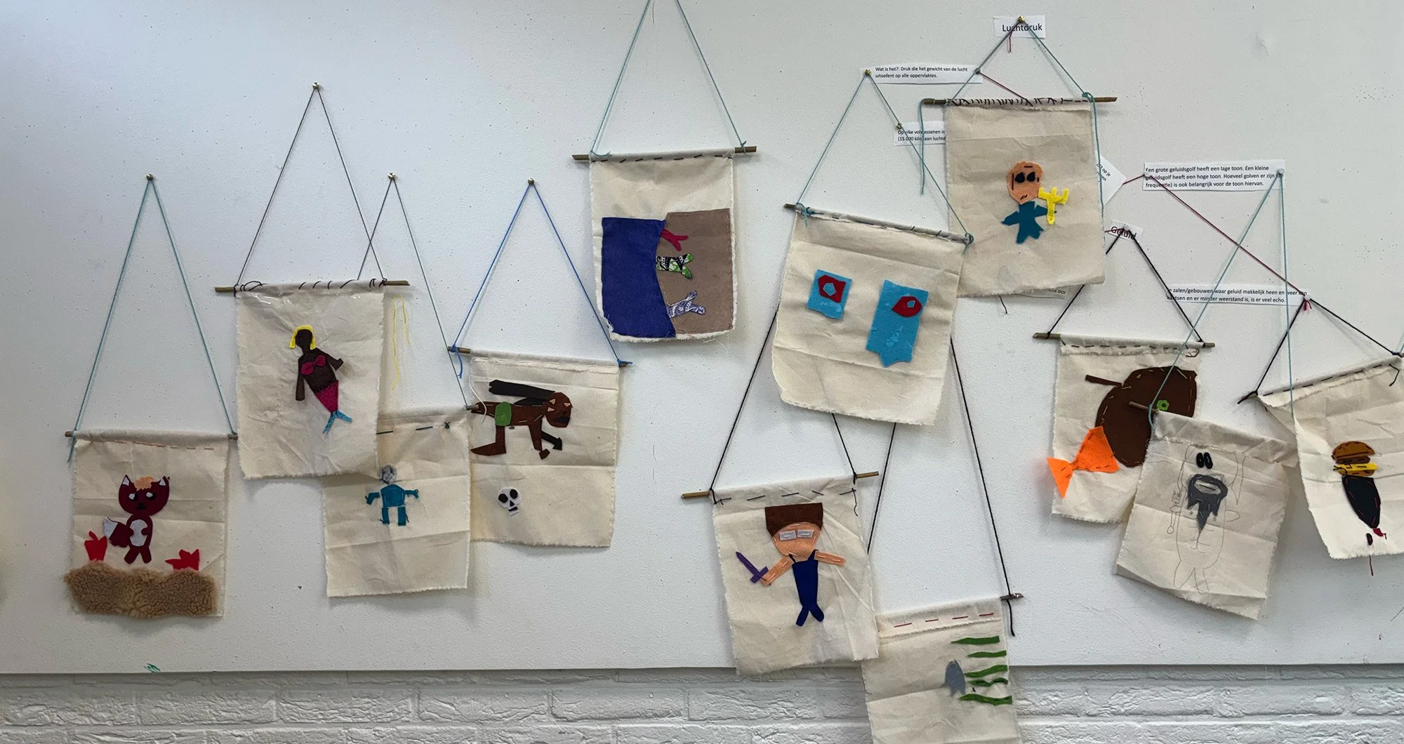 Wall display with multiple small fabric and paper craft artworks, including characters and animals, attached to a string with clothespins, against a white wall.
