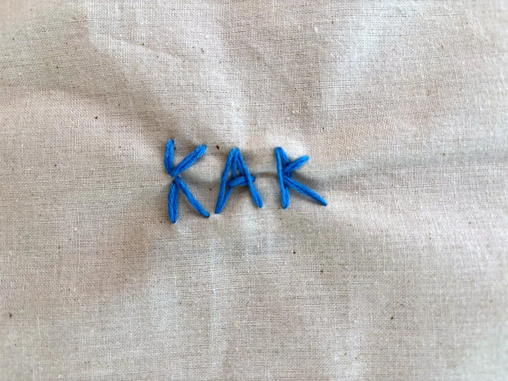 The word "KAK" spelled out with blue embroidery thread on a beige fabric background.