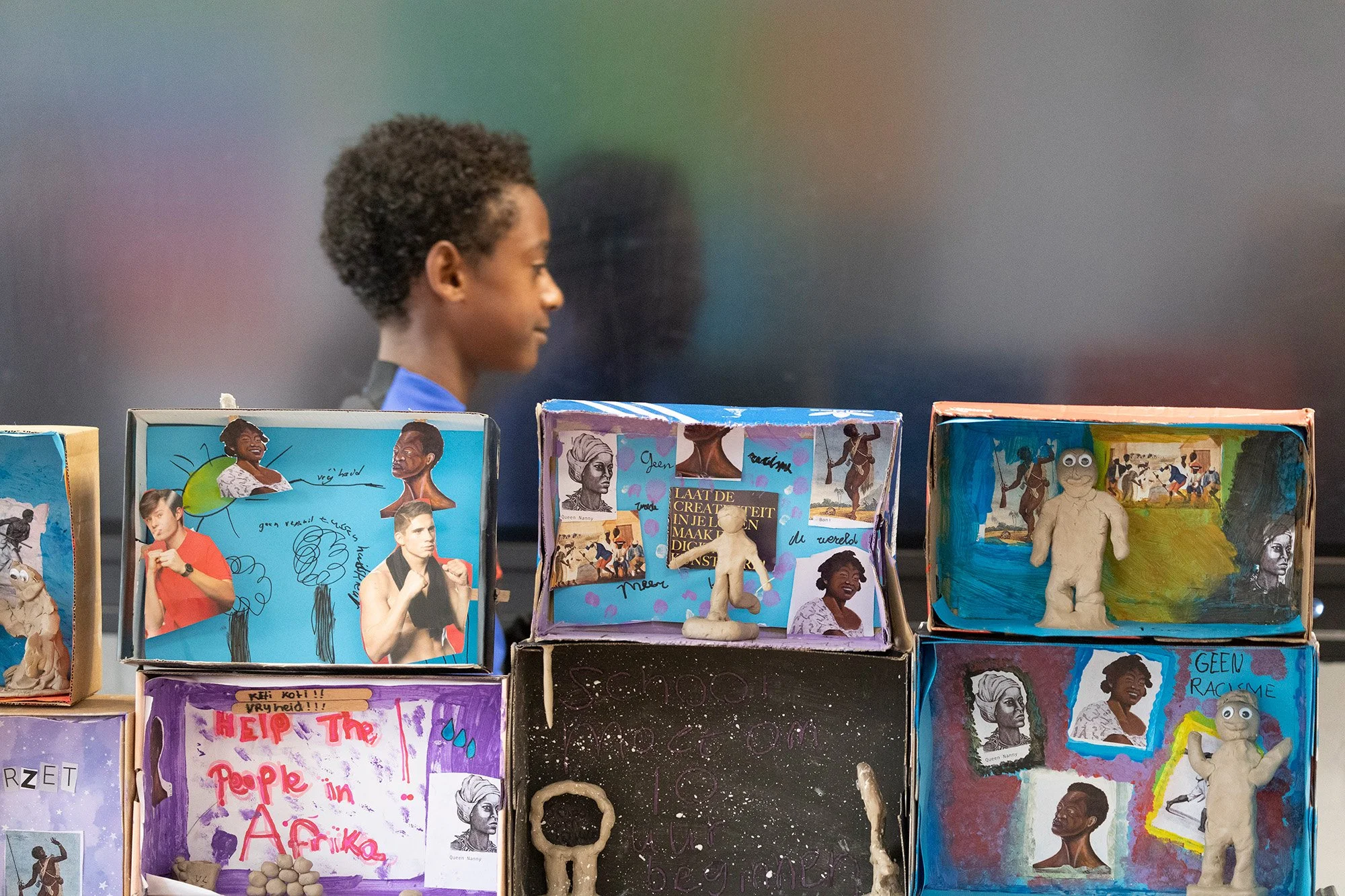 Side view of a boy with curly hair looking at a display of colorful dioramas and standing behind colorful, handmade protest art projects depicting diverse people and messages supporting racial equality.