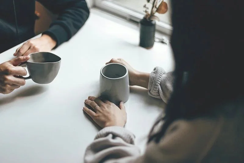 Zwei Personen tauschen ihre Kaffeetassen an einem Tisch in einem modern eingerichteten Raum aus.