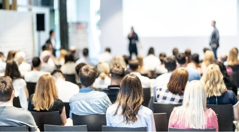 Menschen sitzen in einem Konferenzsaal und hören zwei Sprecher an der Bühne zu.