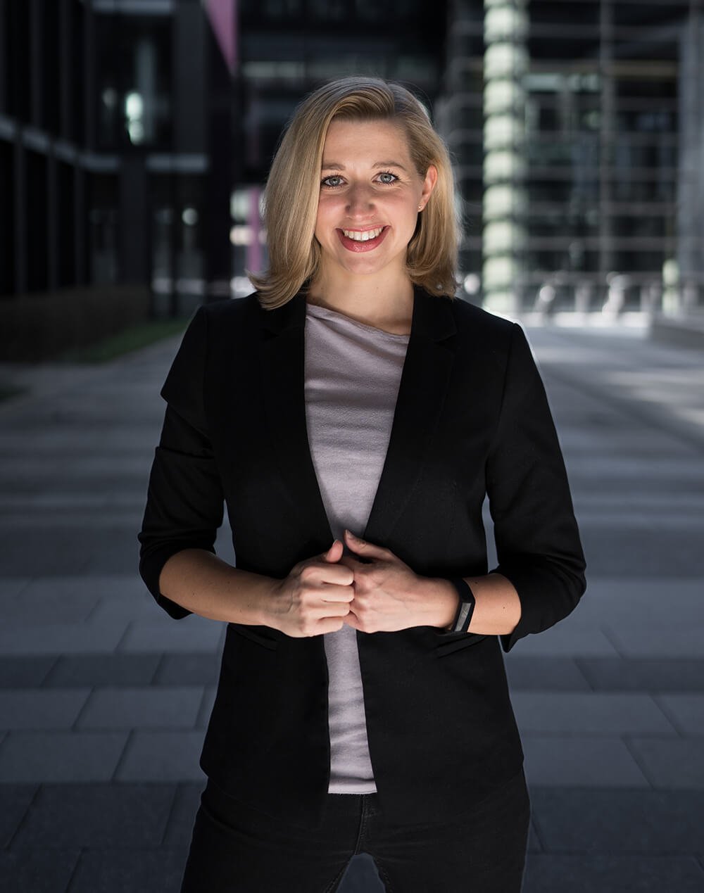 Junge blonde Frau in dunkler Jacke vor modernen Bürogebäuden in der Stadt, lächelnd in die Kamera schauend.