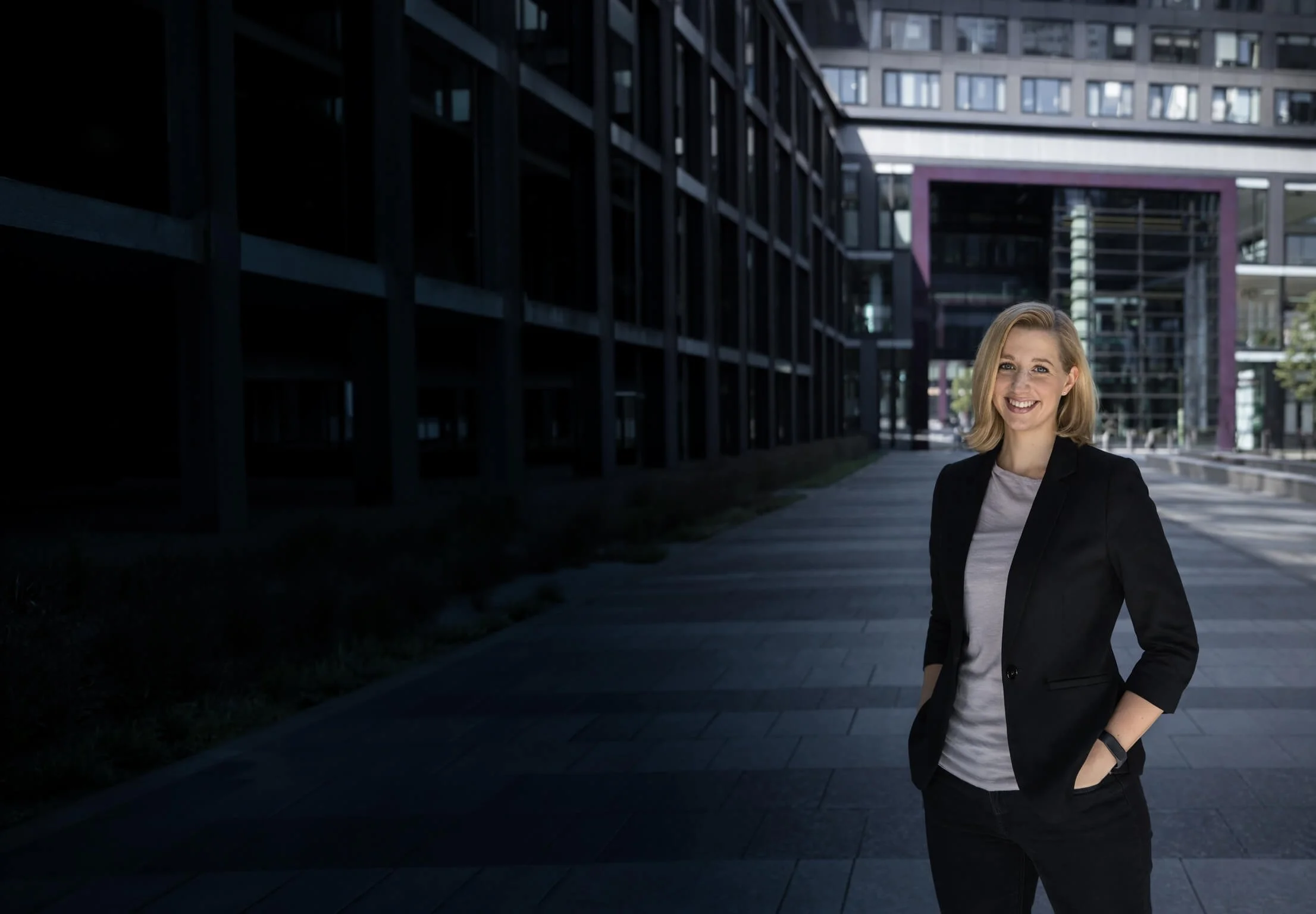 Lächelnde Frau mit blonden Haaren in dunkler Jacke und weißem T-Shirt, stehend vor einem modernen Gebäude mit Glasfassade in einer städtischen Umgebung.